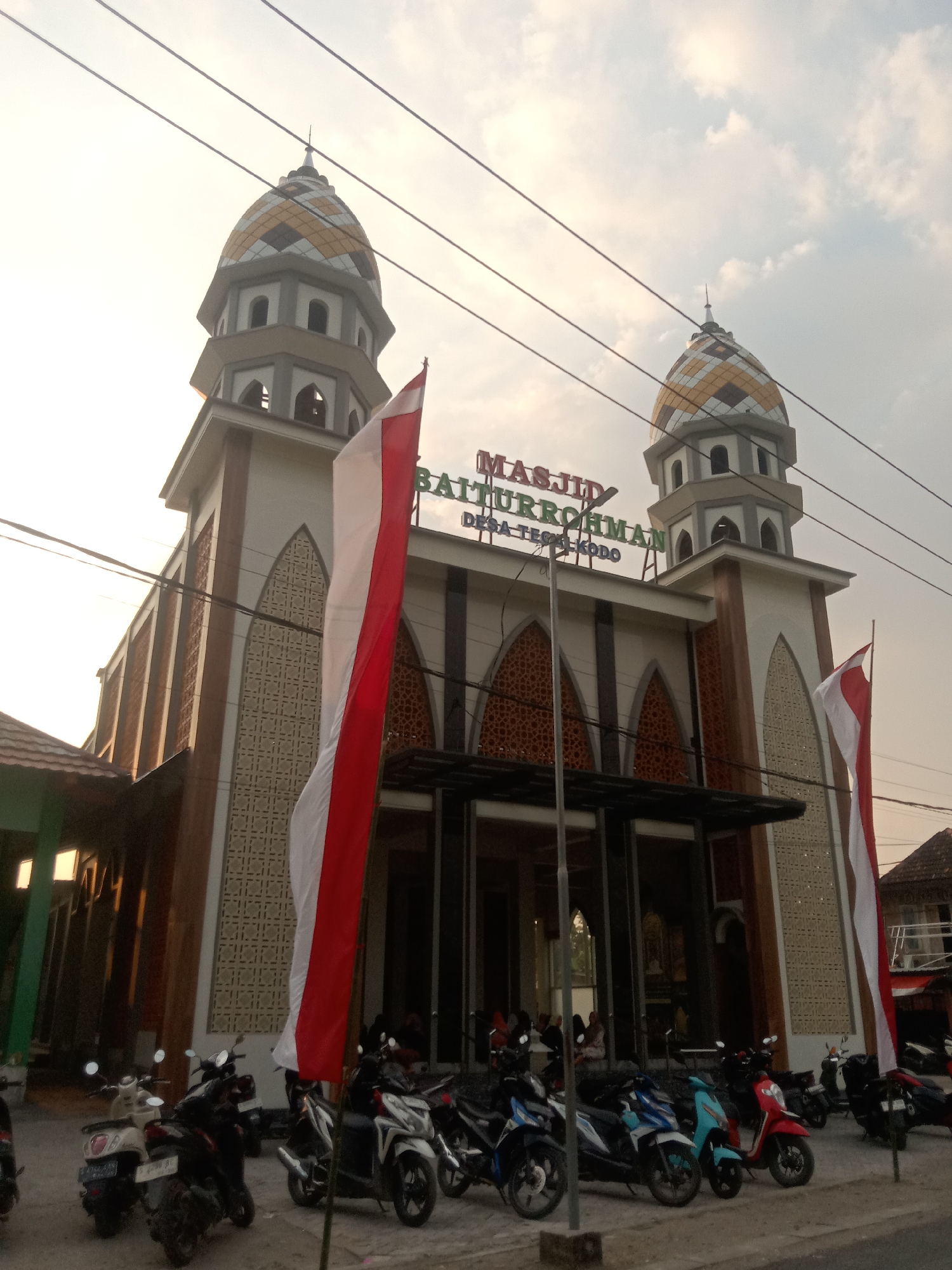 The image shows the exterior of a mosque with two minarets, both topped with patterned domes. The mosque has a modern design with a facade of beige and brown tiles, and large glass doors at the entrance. An Indonesian flag, red and white, is prominently displayed on the right side of the mosque. Several motorcycles are parked in front of the mosque. The sky is overcast with some clouds. The overall lighting suggests it is daytime, possibly late afternoon. The text on the facade reads "MASJID BAITTURROHMAN DESA TEGALKODO".