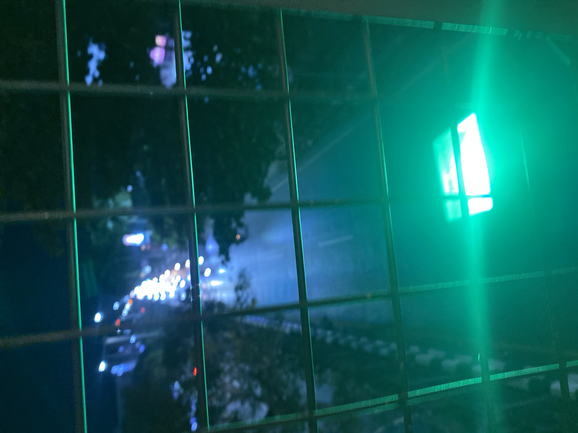 The image depicts a nighttime scene viewed through a metal grate, with a prominent green traffic light dominating the right side. The traffic light is intensely bright, casting a green glow that permeates the scene and creates a lens flare effect. The grate, composed of dark metal bars forming a grid, obstructs the view of the scene beyond, suggesting the vantage point is from behind a fence or barrier.
Through the grate, a road can be seen with blurred, out-of-focus lights of vehicles suggesting traffic. The overall impression is of a city street at night, possibly during a period of moderate to heavy traffic, given the density of lights. The blue and purple hues in the background, along with scattered white and orange lights, hint at streetlights and vehicle headlights/taillights. The presence of trees along the road is discernible, their dark silhouettes softened by the low light conditions.
The time of day is clearly night due to the darkness and the artificial lights. The weather is not explicitly indicated, but the overall ambiance suggests a clear night, as no rain or fog is evident. The location is specified as Banjarbaru, Indonesia, which provides a geographical context to the urban scene. There is no visible text in the image. The emotion evoked is somewhat mysterious and perhaps a little isolating, given the obstruction of the grate and the artificial, somewhat surreal lighting from the traffic signal.
