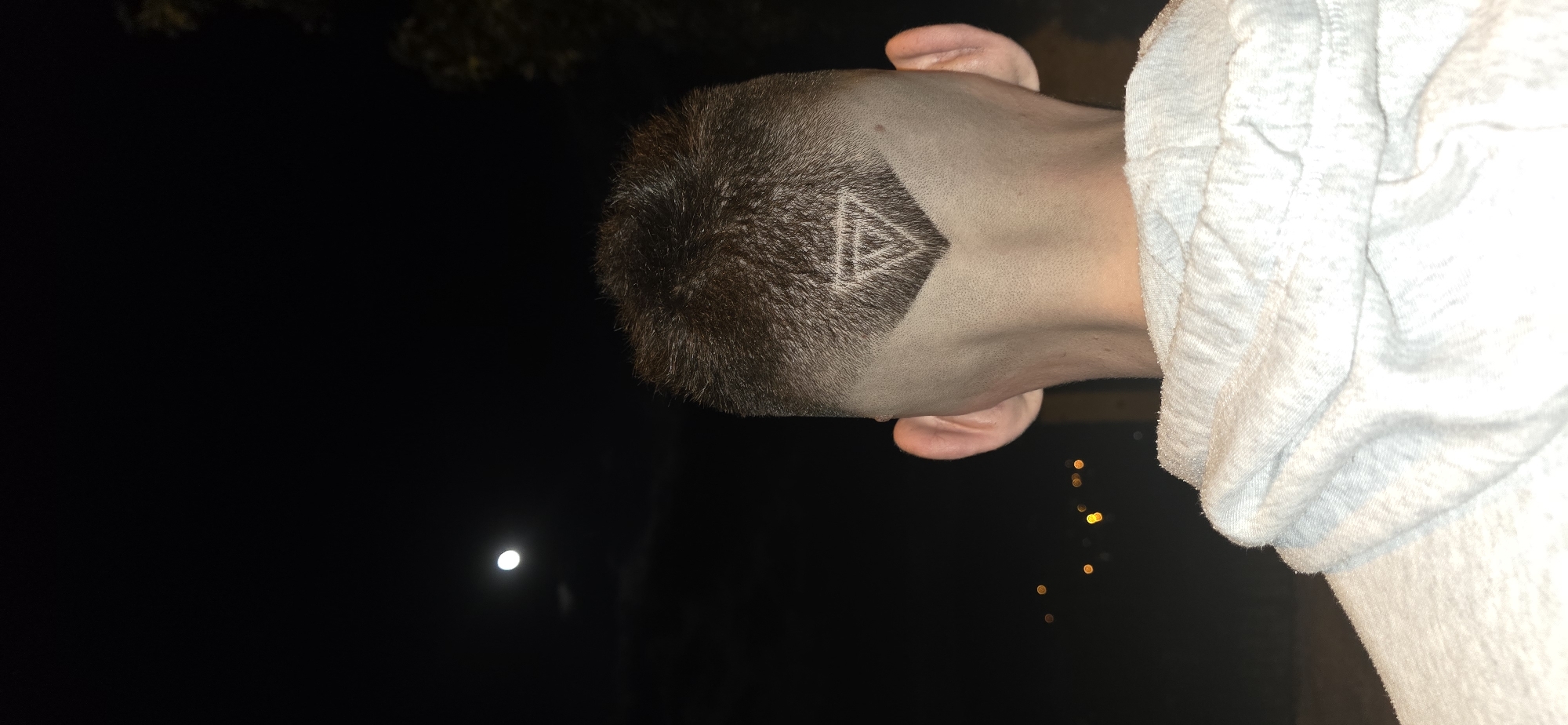 A close-up, slightly angled, and rotated image shows the back of a person's head with a distinctive haircut. The hair is short, dark brown, with a shaved section at the nape of the neck forming a diamond shape. Within the diamond shape, a stylized symbol resembling nested triangles is visible. The person's ears are visible on either side of the head. They are wearing a light-colored, possibly grey or white, hoodie. The background is mostly dark, suggesting nighttime, with a bright light source (possibly the moon) visible in the distance. There are also blurred points of light, possibly streetlights or building lights, in the background on one side. This scene does not contain any information related to the city of Ledenik, Bulgaria.