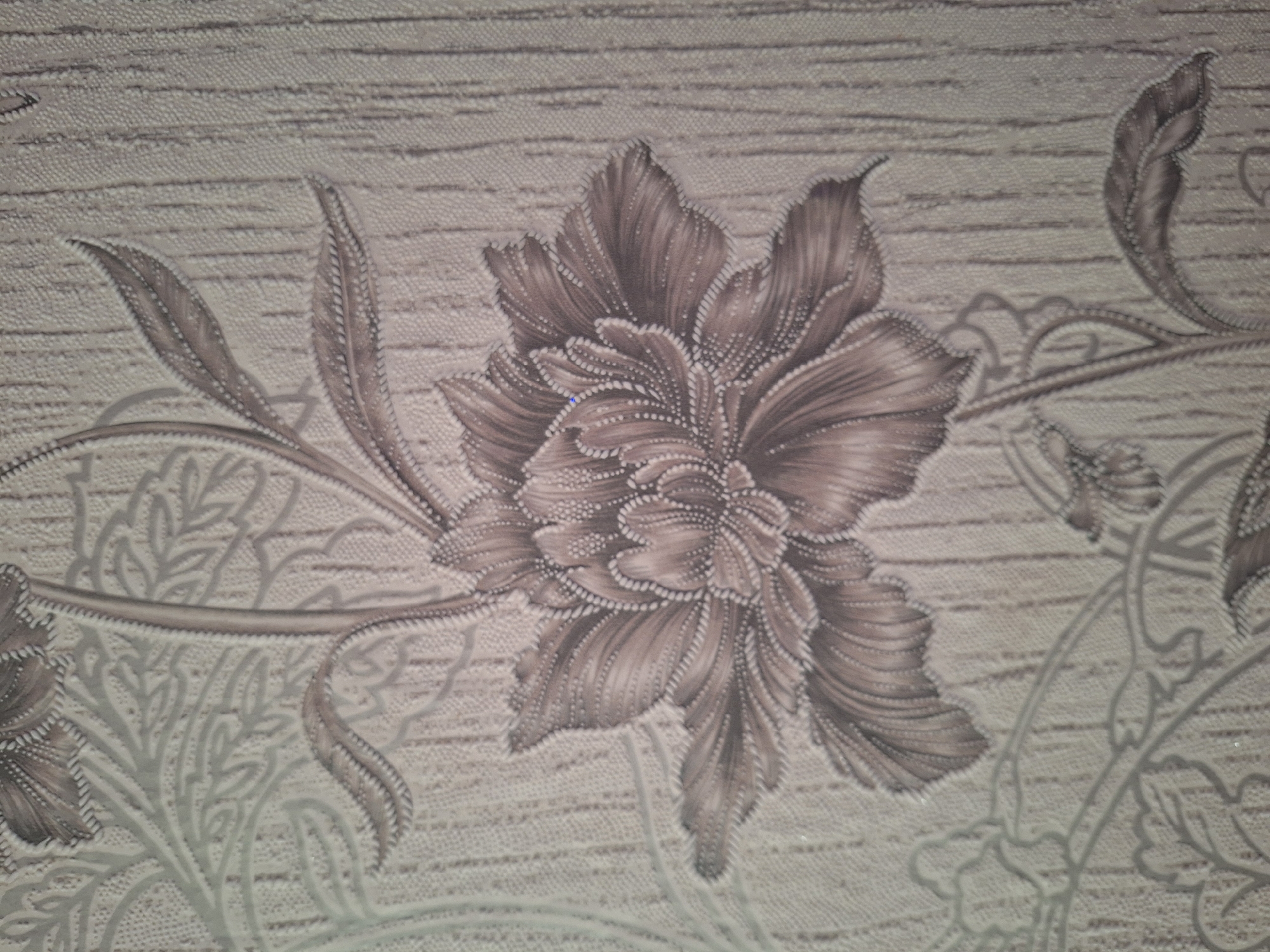 The image shows a close-up of floral wallpaper with a textured, wood-grain-like background. The wallpaper features a prominent, stylized flower with detailed petals in shades of brown and gray. The flower's outline is accented with a dotted pattern. Smaller floral elements and leaves are visible in the background and framing the central flower, rendered in lighter gray tones. The overall impression is one of an interior decorative element. This image does not contain any information that would allow for its specific location within the city of Varna, Bulgaria, or any other geographic area.