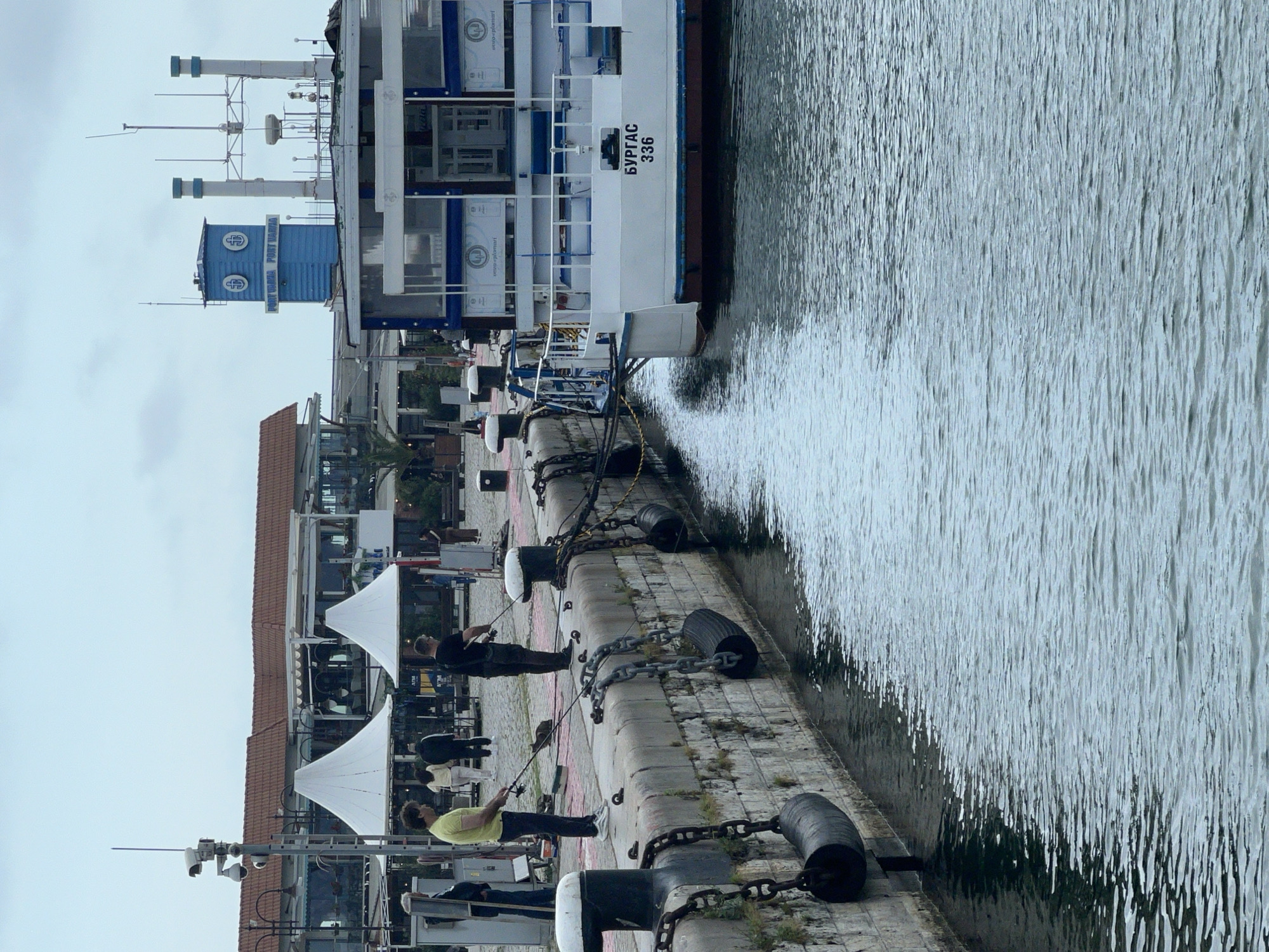 The image depicts a peaceful scene at a port in Varna, Bulgaria, under an overcast sky. A long concrete dock extends into the frame, with rippling water alongside it.
A large white vessel with blue trim is moored at the dock. Its hull clearly displays the Cyrillic text "БУРГАС 336" (Burgas 336). On the boat or an adjacent structure, a distinctive blue building or cabinet features the English text "PORT VARNA" and likely its Cyrillic equivalent, along with a circular logo that appears to be a stylized anchor or "P V" monogram. Several antennas and other maritime equipment are visible on the upper deck of the vessel.
Along the concrete dock, several people are engaged in leisure. Two individuals are prominently seen fishing with rods cast into the water. Other figures are visible further along the pier, some standing near two white, pointed canopies. The dock is equipped with robust black mooring bollards, some linked by heavy chains and incorporating large tire fenders.
In the background, behind the dock, a series of buildings with reddish-brown tiled roofs can be observed, suggesting a waterfront development or promenade. Security cameras mounted on poles are also visible, indicating oversight of the port area. The overall impression is one of calm activity at a working yet recreational port.