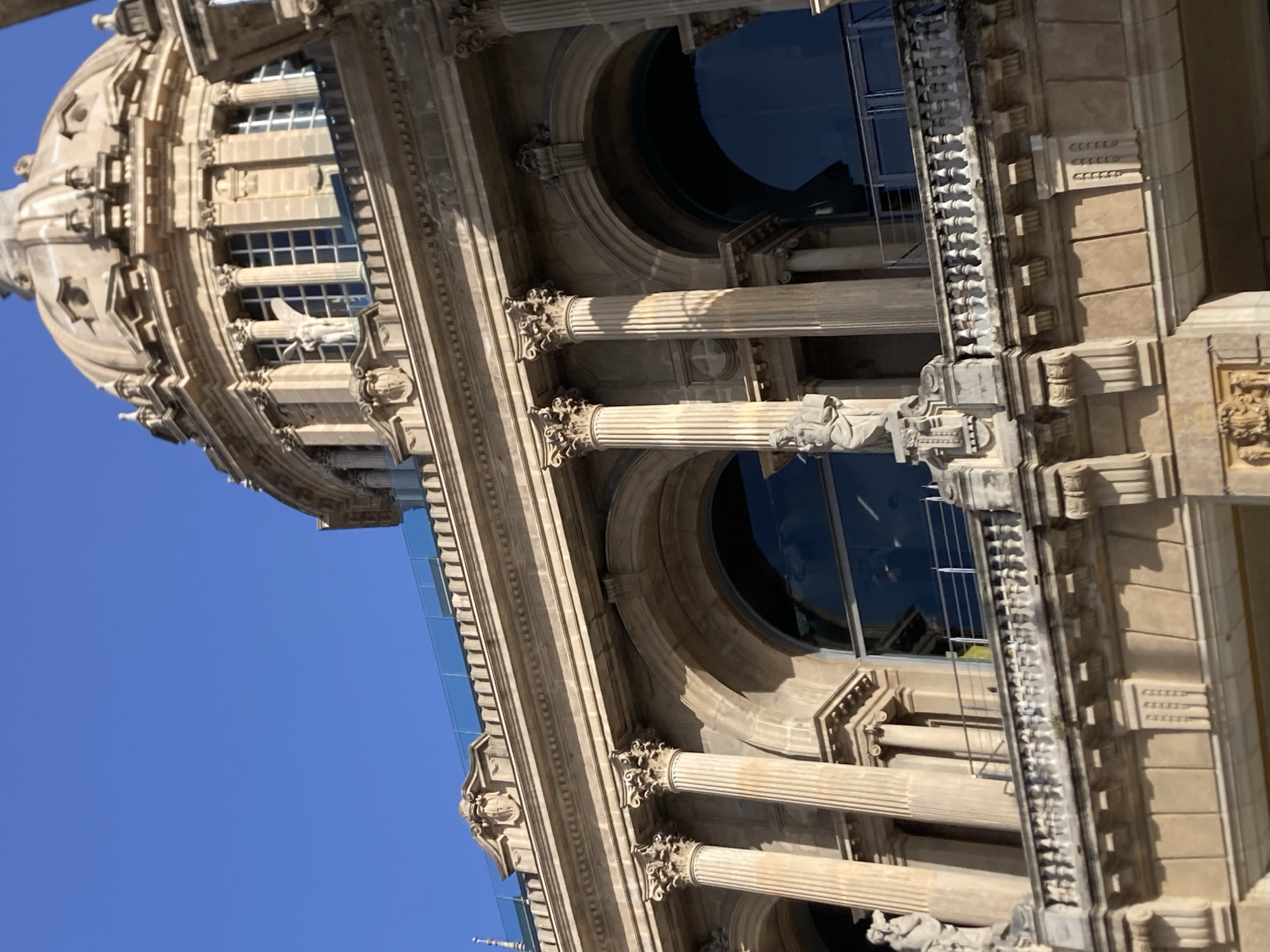 The image shows a close-up of the Palau Nacional in Barcelona, Spain, with its distinct dome and classical architecture. The building's façade is made of stone and features arched windows, Corinthian columns, and ornate detailing. A balcony with a white railing runs across the middle of the structure, and a statue of a winged figure can be seen on the left side of the building. The sky is a clear blue, suggesting a sunny day. The perspective is from a low angle, looking up at the building.