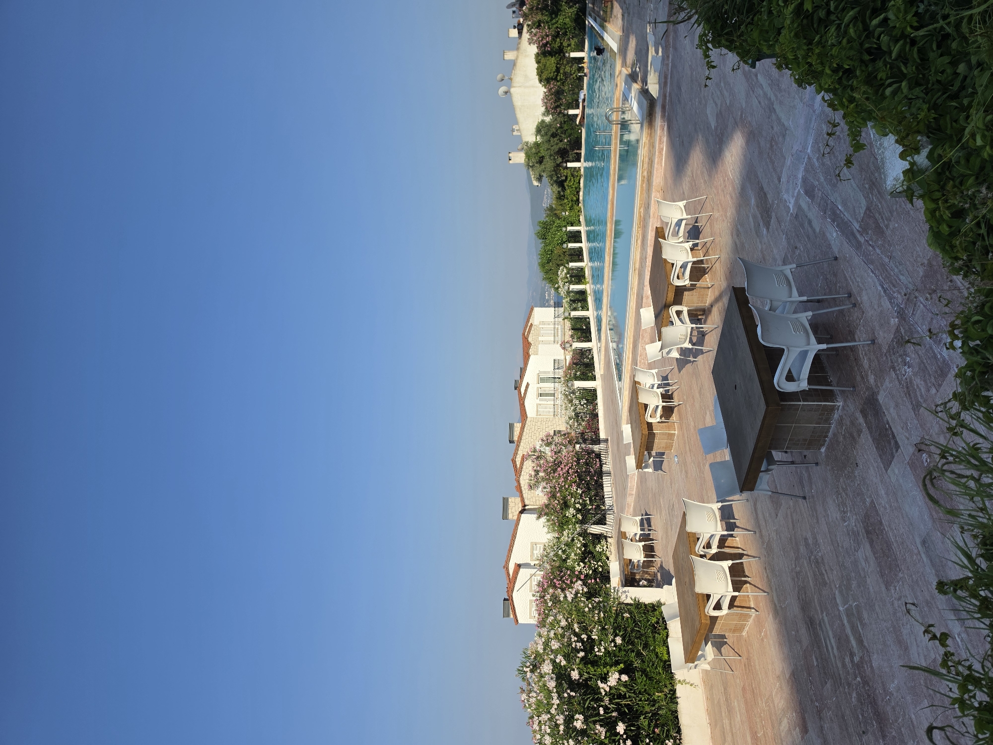 The image captures a serene outdoor scene in Urla, Turkey, featuring a swimming pool area with seating arrangements. The foreground is dominated by a spacious patio paved with light-colored, marbled tiles, on which several wooden tables are placed, each surrounded by white plastic chairs. Some tables are set for dining, while others appear to be unoccupied. Lush green bushes and flowering plants frame the patio on the right and left sides.
Beyond the patio, a rectangular swimming pool with clear blue water is visible. The pool has steps for entry and appears to be moderately occupied with a few individuals swimming or relaxing. The area surrounding the pool is also paved.
In the background, a row of houses with white stucco walls and red-tiled roofs are situated. These houses are adorned with stone accents and chimneys, suggesting a residential or resort-like setting. Some of these houses are partially obscured by vibrant green foliage and flowering bushes, predominantly pink and white oleander. The sky above is a clear, unblemished blue, indicating fair weather. The overall impression is one of a tranquil, well-maintained leisure space in a coastal or hilly area.