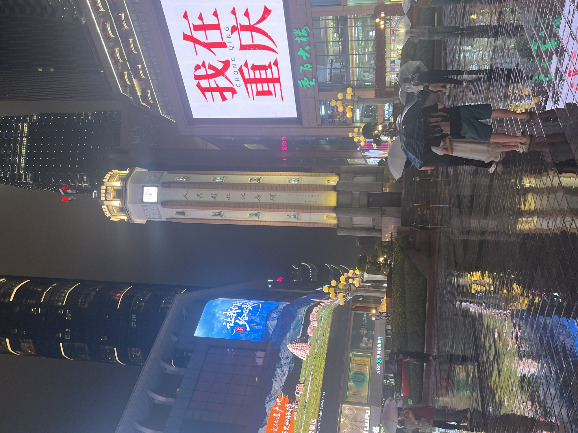 At night in Yuzhong District, China, a group of people stand on a wet, tiled plaza, many holding umbrellas. The plaza reflects the neon lights and signage from surrounding modern buildings, including a large digital billboard displaying Chinese characters in red and white, with "CHONG QING" in smaller English text below. To the left, a tall skyscraper with illuminated horizontal bands dominates the skyline, partially obscuring another modern building. In the center of the frame, a multi-story tower with a clock face stands prominently, adorned with vertical text in Chinese characters. Further to the right, another building features illuminated balconies and a prominent sign with green Chinese characters. The overall atmosphere is urban and vibrant, despite the rainy conditions.