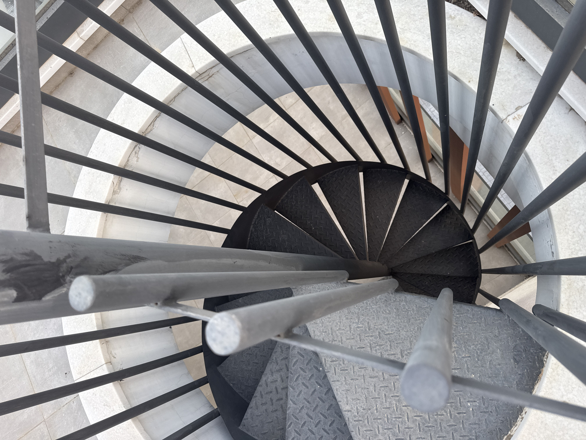 A top-down, close-up view captures a spiral staircase made of metal. The staircase features diamond plate treads, with some sections appearing darker than others, possibly due to wear or different materials. A central metal pole supports the structure, with horizontal railings made of vertical metal bars forming a circular enclosure around the staircase. The surrounding area shows light-colored tiling with some visible grout lines. The lighting suggests an outdoor environment. The image is taken in Agios Athanasios Municipality, Cyprus.
