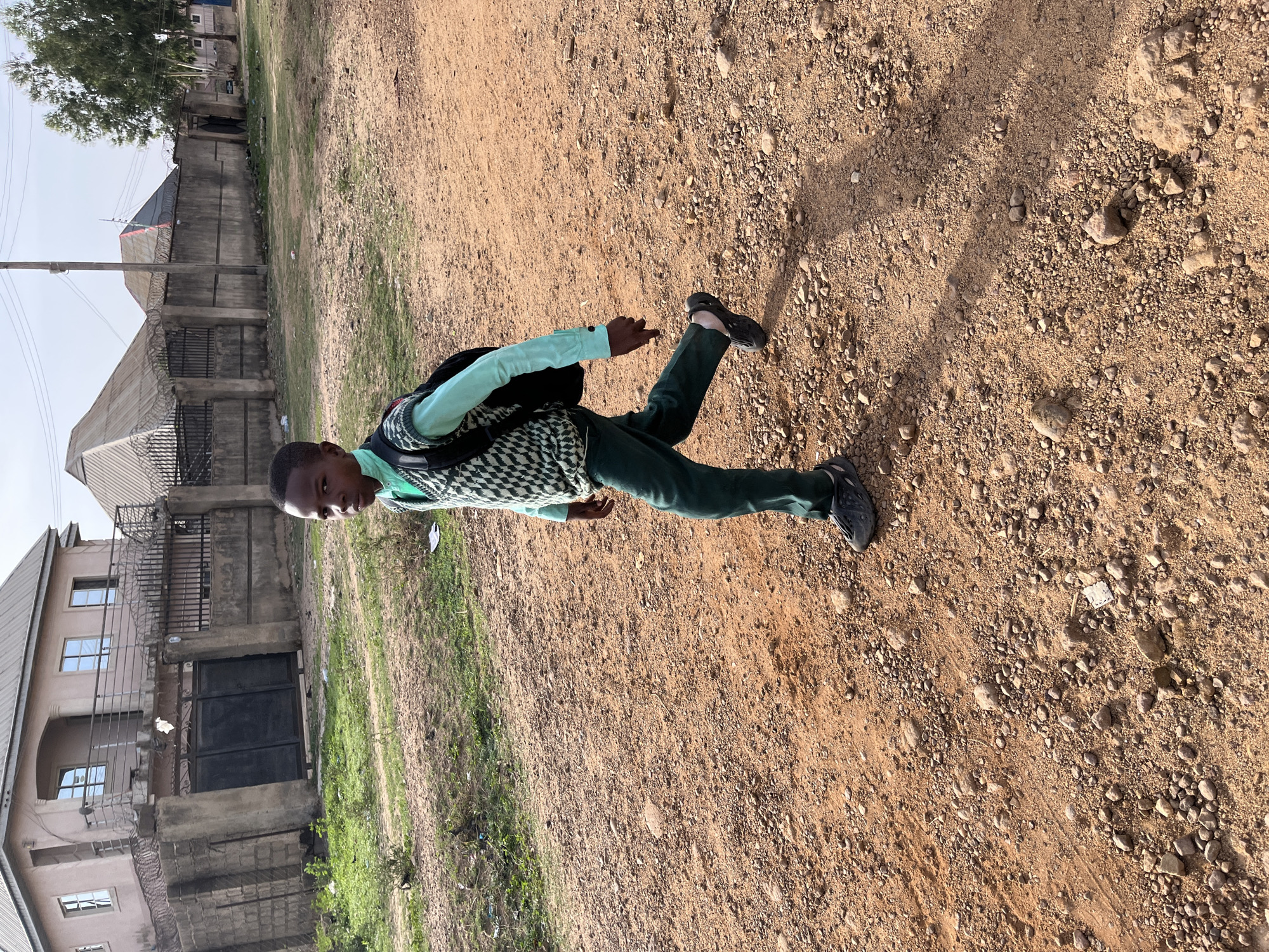 A young person, seemingly male, is walking on a dirt or gravel path. The young person is wearing dark pants and shoes, and a patterned shirt or sweater, and carrying a backpack. The background includes buildings and vegetation. The scene appears to be outdoors. It is difficult to pinpoint the exact location in Jos, Nigeria, without further details, but based on the environment, it could be a residential area.