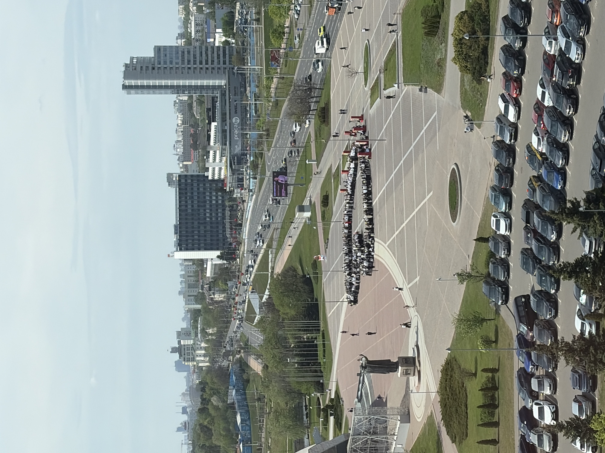 The image shows an aerial view of a city, likely Бараўлянскі сельскі Савет, Belarus, with a wide boulevard featuring significant traffic. A large, paved plaza dominates the foreground, containing a monument and people gathered in rows, possibly for an event. A parking area is visible with many vehicles. The environment appears to be urban, with buildings, trees, and green spaces.