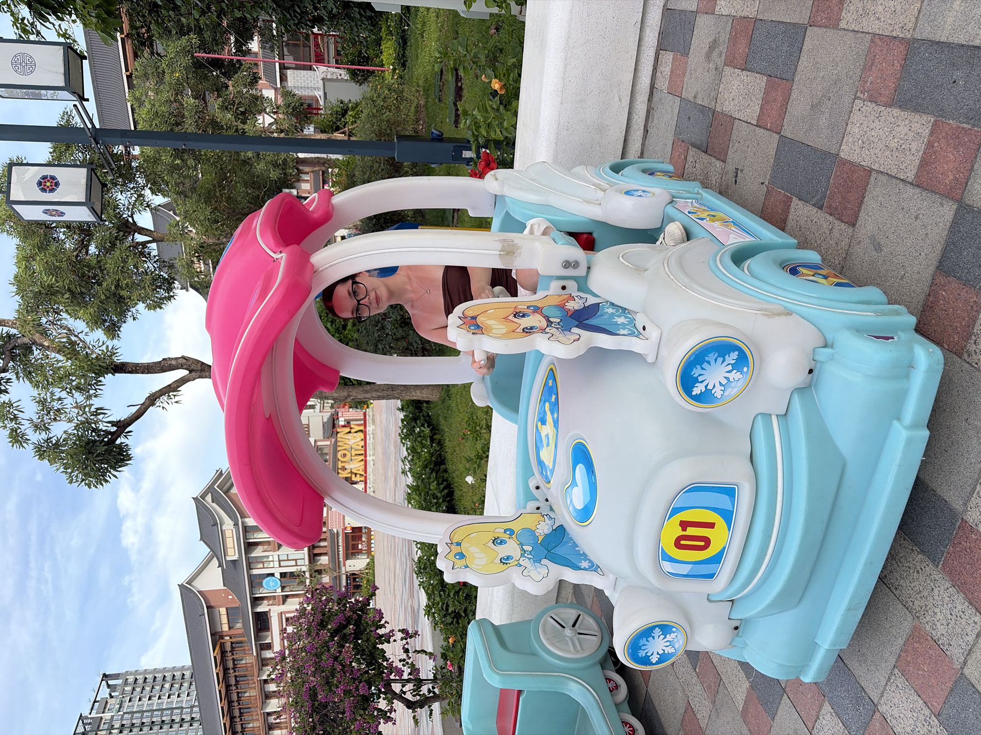 A young woman wearing glasses and a brown strapless top sits in the driver's seat of a light blue and white toy car with a pink canopy. The car is decorated with cartoon princess figures, snowflakes, and a yellow oval with the number "01" inside. She is holding the steering wheel, and her body is positioned as if she is driving. The car is on a paved area with a patterned stone surface. In the background, there are buildings with traditional Asian-inspired architecture, trees, and a tall modern building. The sky is partly cloudy. The environment appears to be a public outdoor space, possibly a park or a recreational area.