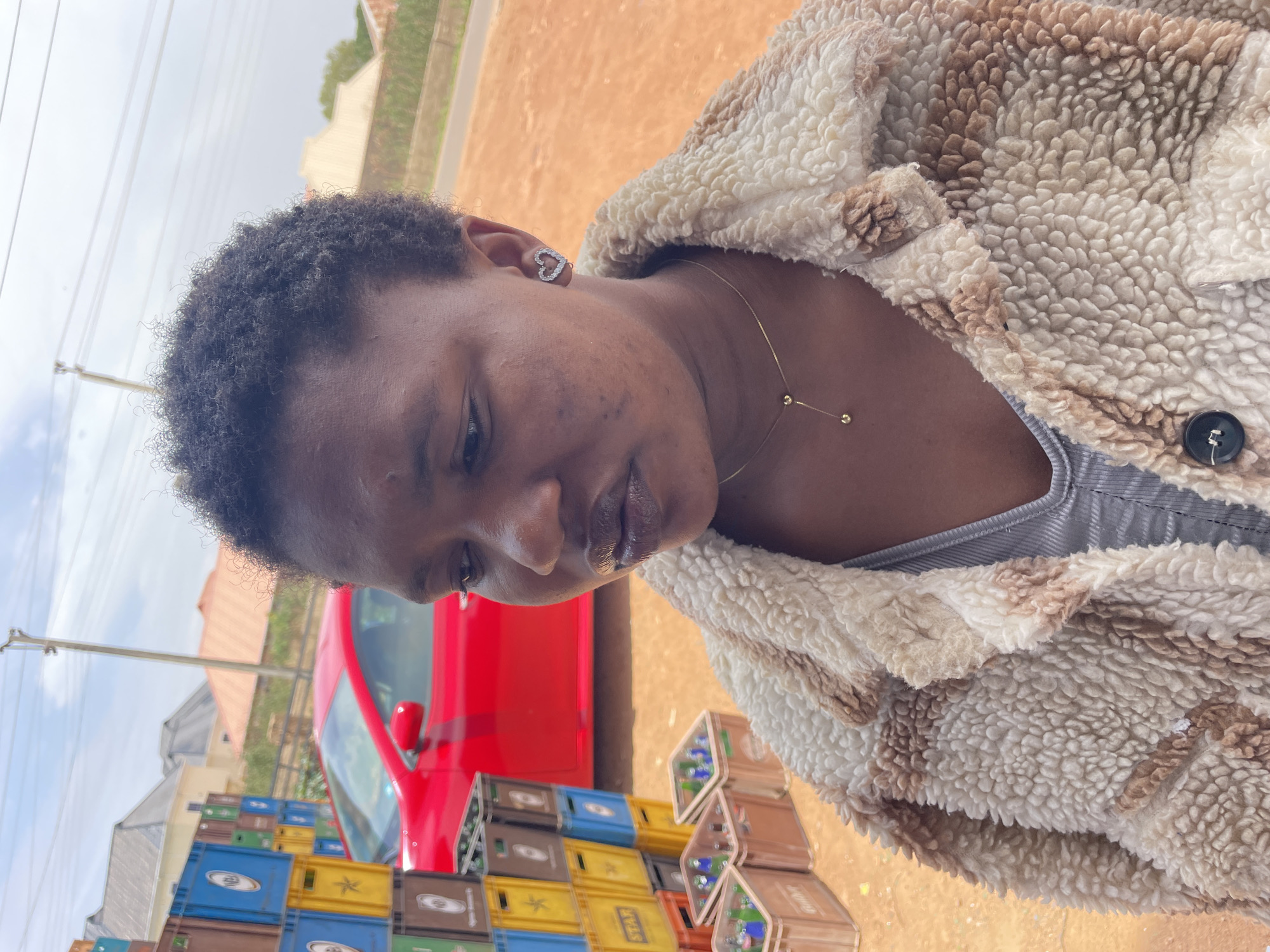 A person with short, dark curly hair, light brown skin, wearing a furry brown and white coat and a light gray shirt, stands near a stack of colorful plastic crates containing glass bottles, next to a red car. The location appears to be outdoors in Gura Top, Nigeria, based on the context of the query. The person is wearing a small heart-shaped earring and a thin gold necklace.