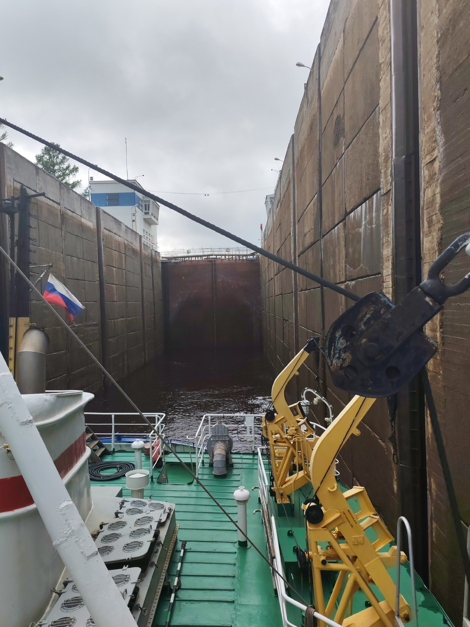 The image shows a view from the deck of a vessel navigating a canal lock. The high concrete walls of the lock are visible on either side. The Russian flag is visible on the port side of the vessel. The vessel is equipped with yellow cranes and other deck machinery. The water level in the lock is below the top of the walls. A control tower is visible in the distance. The image seems to show a boat passing through the Volga–Baltic Waterway near Devyatiny, Russia.