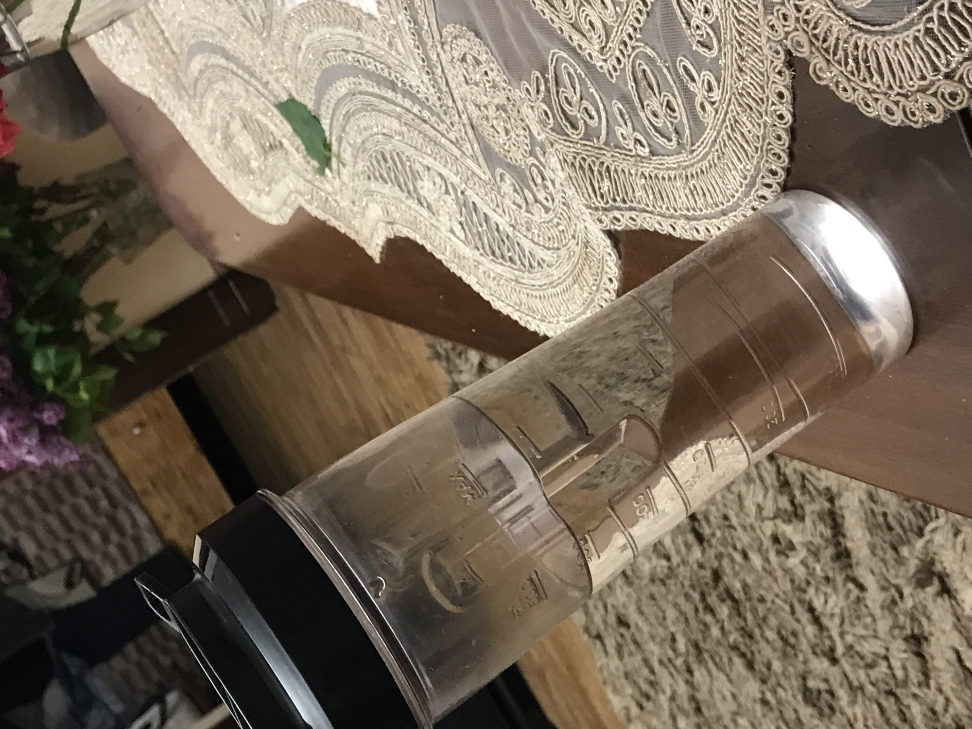 The image shows a plastic blender container with a black base and silver lid lying on a surface with an embroidered lace tablecloth. The background is a blend of wooden furniture and a carpeted floor. The container is transparent, partially filled with a light brown liquid or a powdery substance. There's no indication of the location being Yerevan, Armenia.