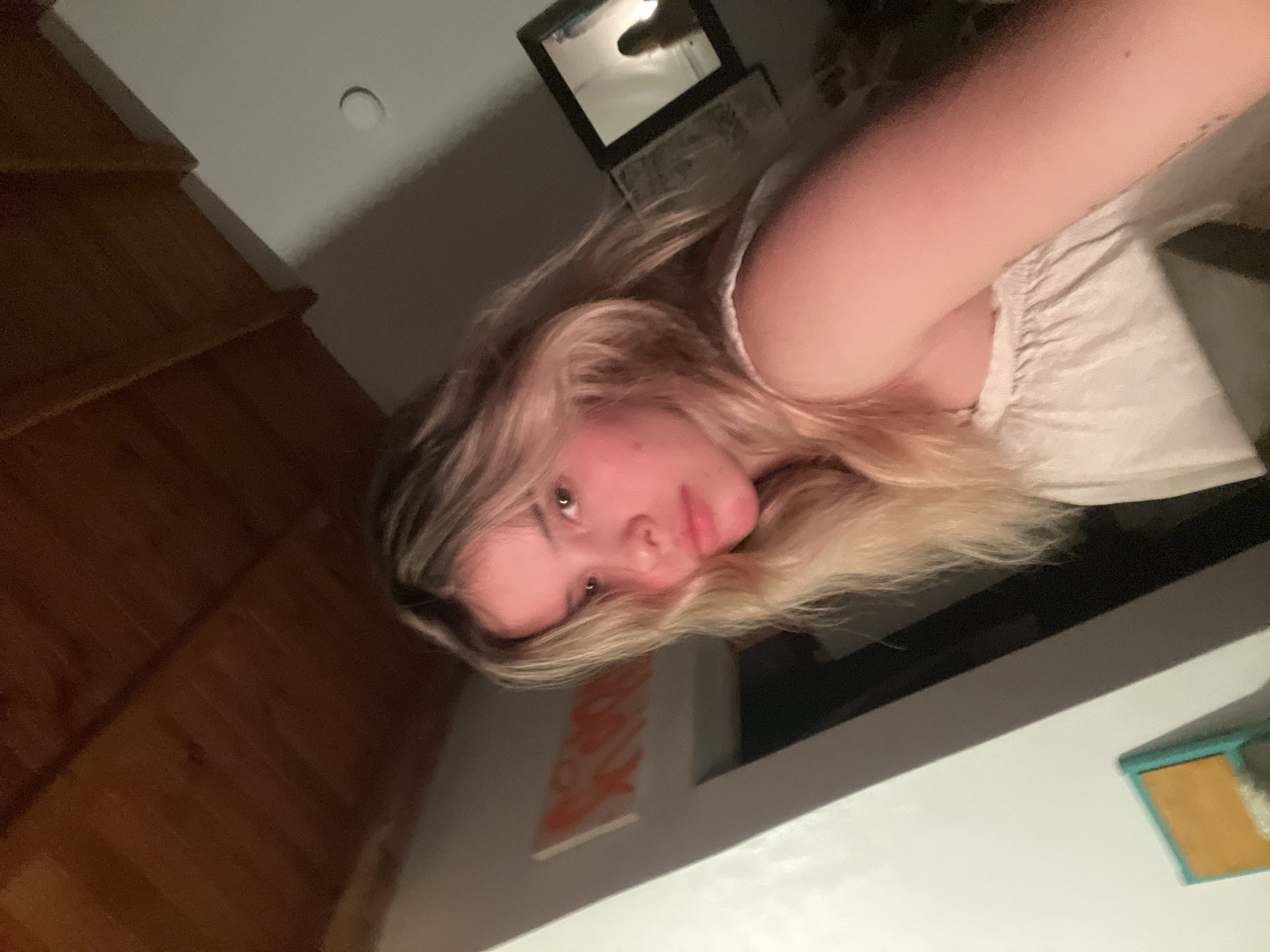 The image shows a young woman with blonde hair looking at the camera. She is wearing a white top. The background is dimly lit and includes a wooden ceiling, a wall with a light fixture, a framed artwork, and a mirror reflecting a silhouette of a person. A small, colorful object is visible in the lower left corner. The scene appears to be indoors. The location is specified as Sumalao, Argentina.