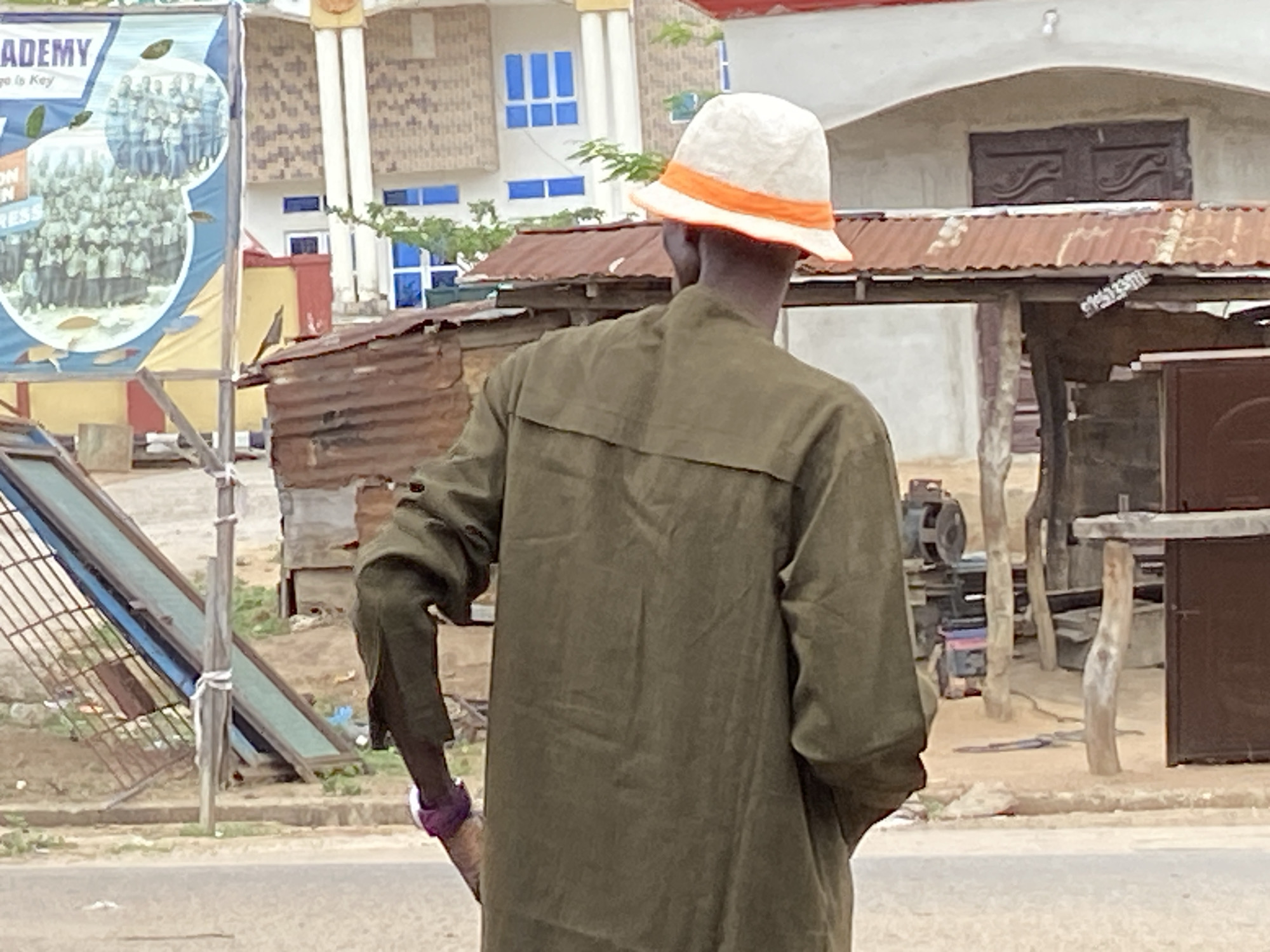 A man wearing a white bucket hat with an orange band and a green, long-sleeved shirt is standing on a street in Mararaba, Nigeria. His back is to the camera, and he is holding something in his left hand. In the background, there is a corrugated metal structure and a building with blue-trimmed windows. A sign with an image of students is visible on the left.