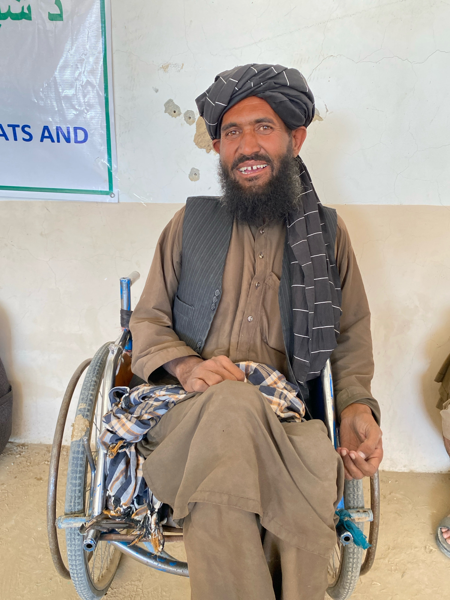 A man with a beard and a turban sits in a wheelchair in an indoor setting. He is wearing a brown robe and a dark vest. A checkered cloth is draped over his lap. The wall behind him is off-white and appears to have some damage, possibly from bullet impacts. To the left of the man, a white banner with green and blue lettering is partially visible, with the words "ATS AND" clearly legible in blue. The floor is a light brown, dusty surface. The scene appears to be in Pasab, Afghanistan.