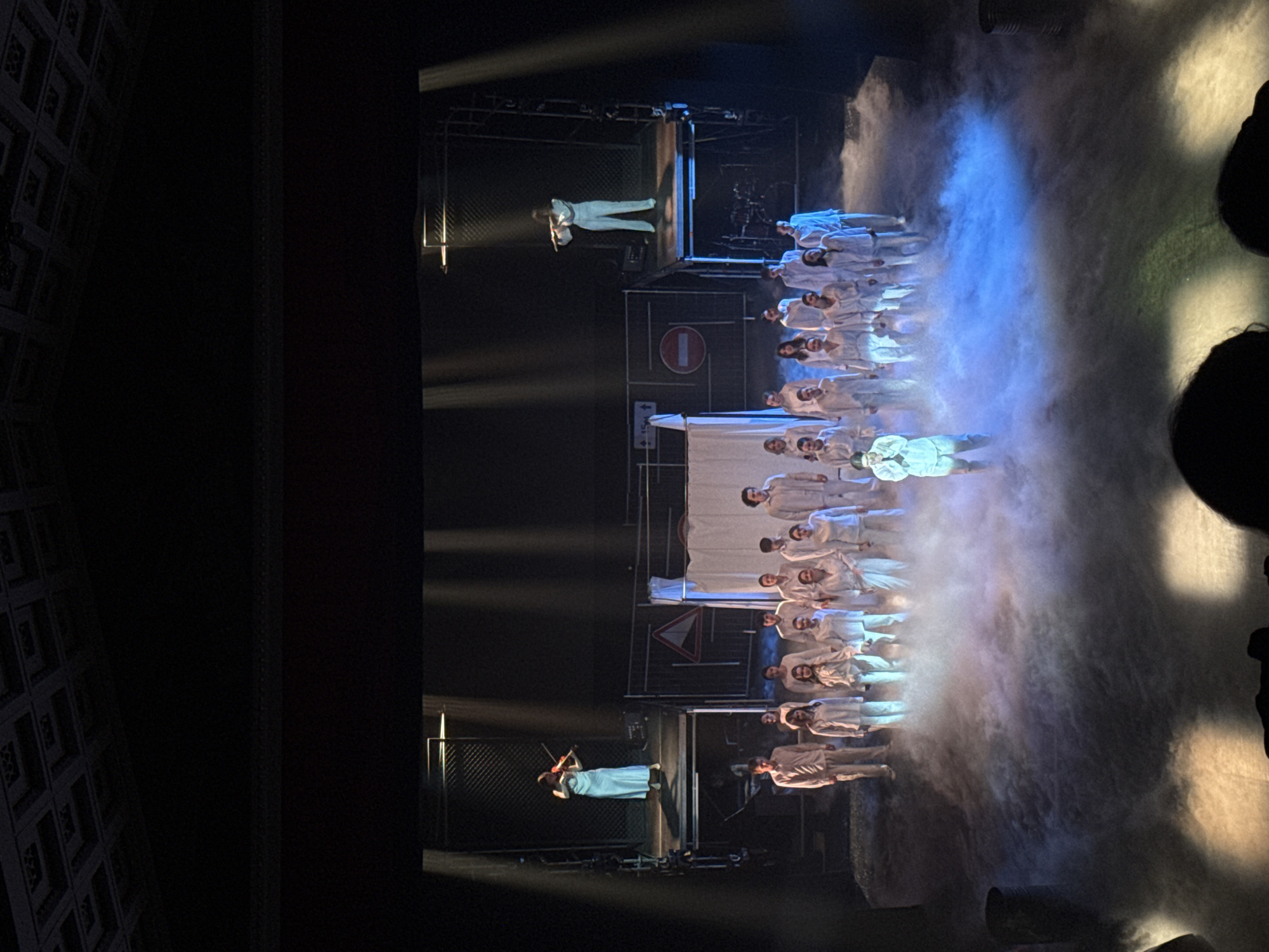 The image shows a stage performance, likely in a theater, with a group of performers dressed in white uniforms. They are illuminated by a combination of white and blue stage lights. The backdrop features metal scaffolding and what appears to be road signs. Two individuals are positioned on elevated platforms, one holding and playing a violin. The environment includes artificial fog or smoke effects. The location is possibly Perm, Russia, due to the cultural performance context.