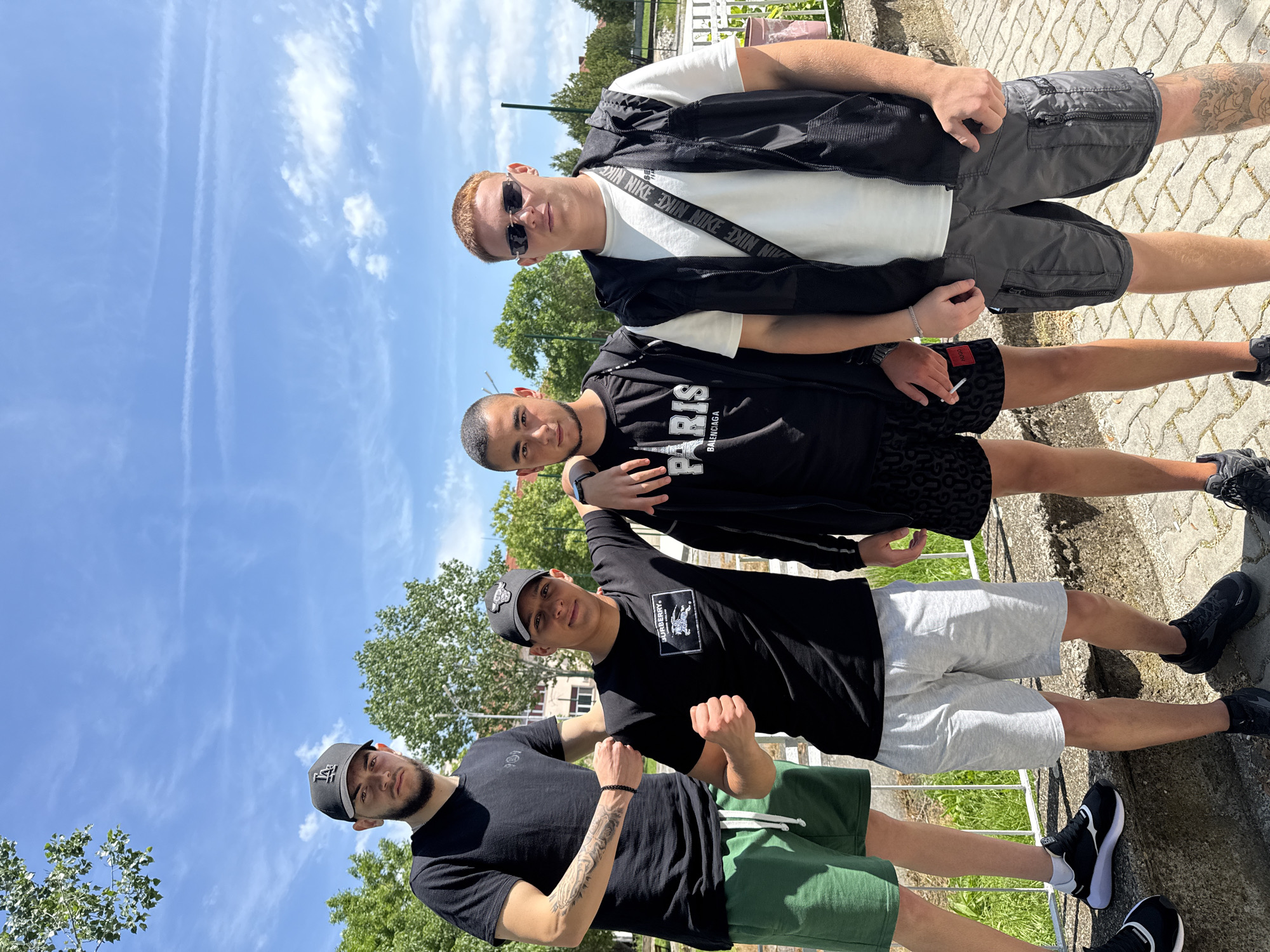 Four young men are standing in a row outdoors. The man at the top is wearing a white t-shirt, grey shorts, a black jacket, and sunglasses. The second man is wearing a black t-shirt with "Paris" on it and black patterned shorts. The third man is wearing a black t-shirt and grey shorts, and he is raising his fists. The bottom man is wearing a black t-shirt and green shorts. They are standing on a paved surface with a fence behind them. The sky is blue with white clouds, and there are trees in the background. This location is in Elhovo, Bulgaria.