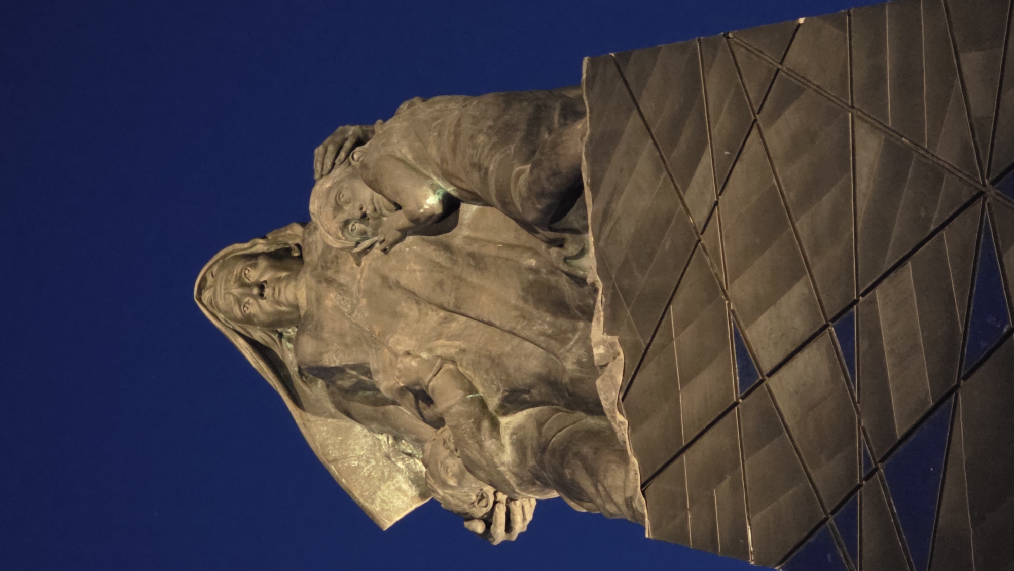 A low-angle shot shows a monument or sculpture, positioned against a solid blue sky. The sculpture depicts a woman holding a child, rendered in a weathered bronze or similar material. To the right of the sculpture is a large, geometric structure, composed of numerous triangular and diamond-shaped panels. The monument is located in Izhora (Verevskoe S/P), Russia.