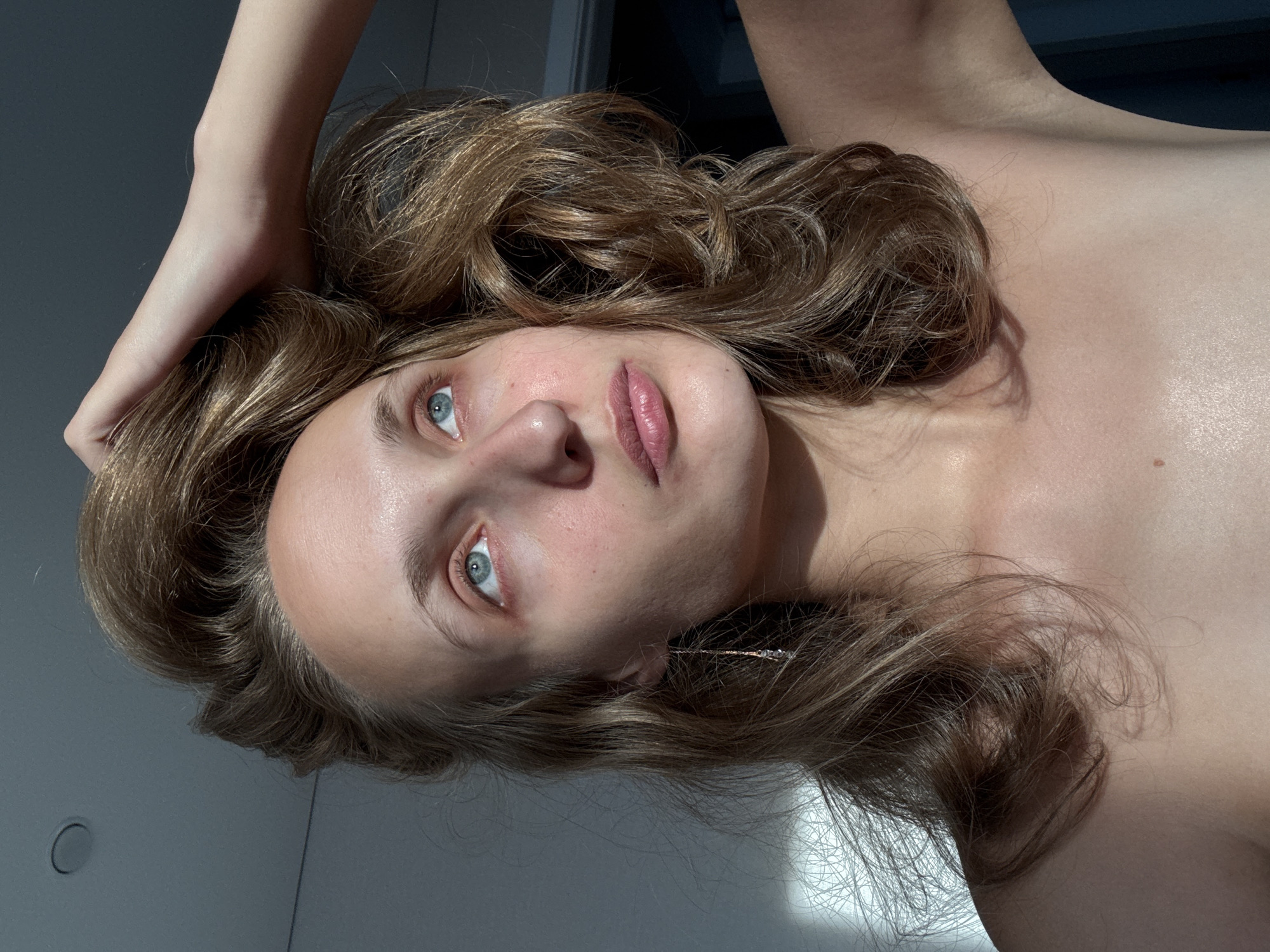 A woman with light brown wavy hair is captured in a close-up portrait, looking upwards and to her right, with her left hand resting on top of her head, playing with her hair. The lighting suggests an indoor environment, possibly a room in Podshivalovo, Russia, with natural light streaming in from the left, casting shadows on the wall and the subject's skin. Her eyes are a striking blue, and her lips are slightly parted. She is wearing a delicate dangling earring on her left ear. The focus is on her face and upper body, with her bare shoulders visible. The background is out of focus, with hints of a ceiling and a dark doorway.