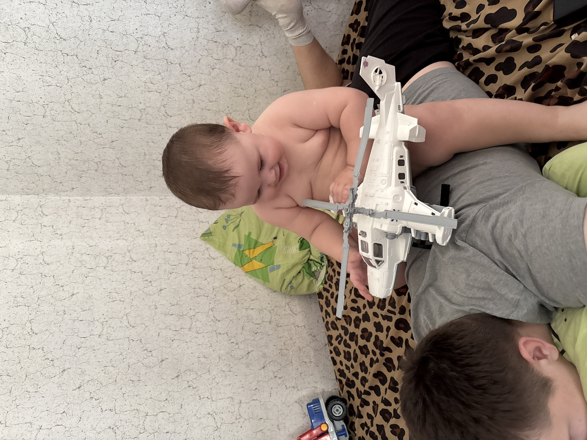 A top-down, indoor shot depicts a baby, approximately 1 year old, lying prone on an adult who is reclining on a bed with a leopard-print cover. The baby, dressed only in a diaper, faces left and is focused on a large white toy helicopter in front of them. The adult, wearing a grey t-shirt and shorts, is partially visible, with the top of their head, ear, and shoulder visible on the right side of the frame. A green patterned pillow is partially visible underneath the baby. In the lower left corner, a partially visible blue and red toy truck sits on the floor against a wall. The precise location in the city of Фоки, Russia, cannot be determined from this image.