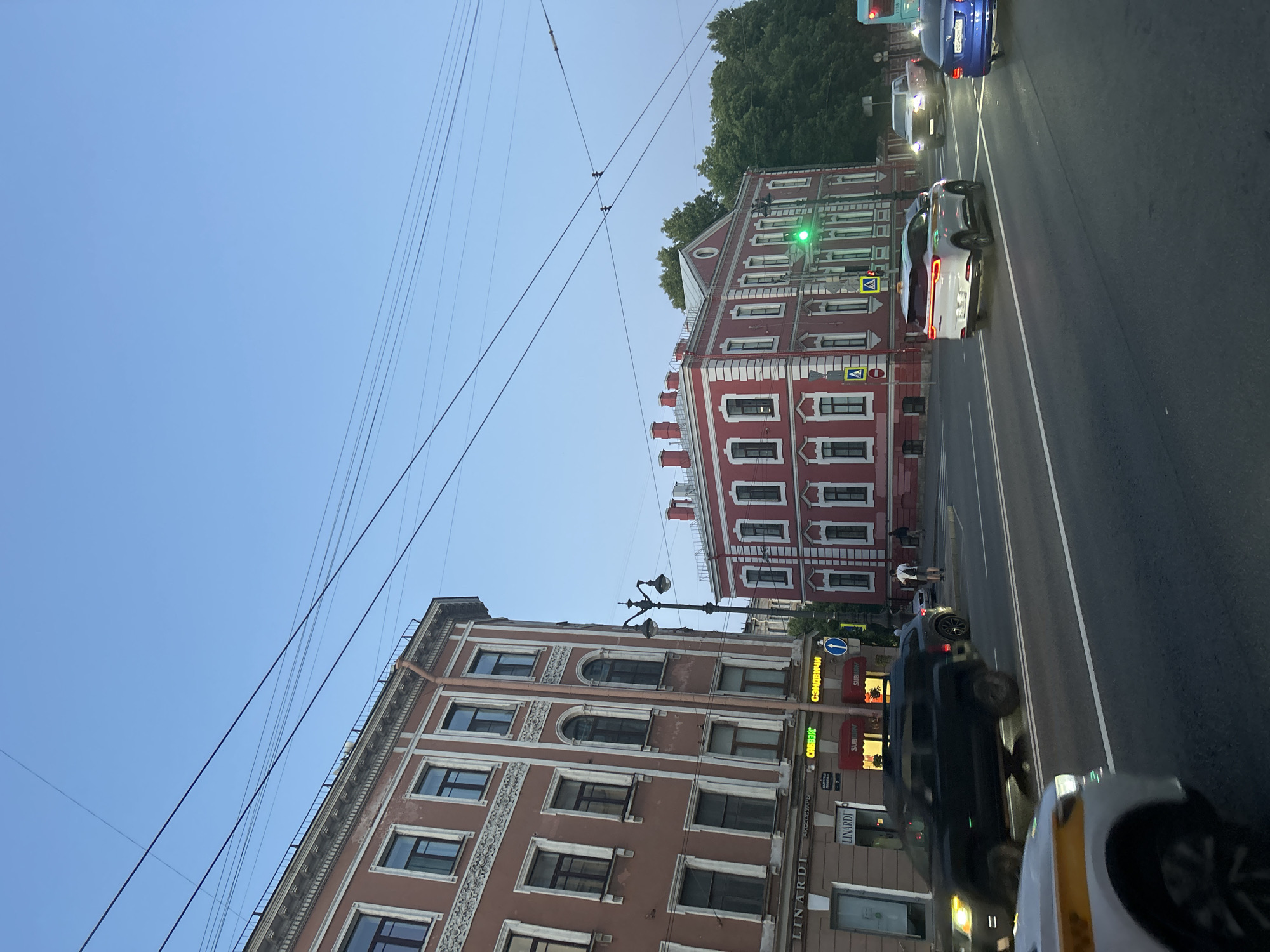 The image shows a street scene in a city, likely during dusk or early evening. A multi-story building with a red facade and white trim dominates the right side of the frame, featuring multiple windows and chimneys. To the left, another large building with a reddish-brown facade and ornate white trim is visible. A street with cars and a traffic light is in the foreground. A green traffic light is illuminated. There are several cars on the road, including a white SUV, a dark SUV, a white car with yellow stripes, and a blue sedan. Trees are visible in the background behind the red building. Power lines crisscross the sky. A few pedestrians are on the sidewalk in the distance. Signs for "LINARDI" and "SUBWAY" are visible on the buildings.