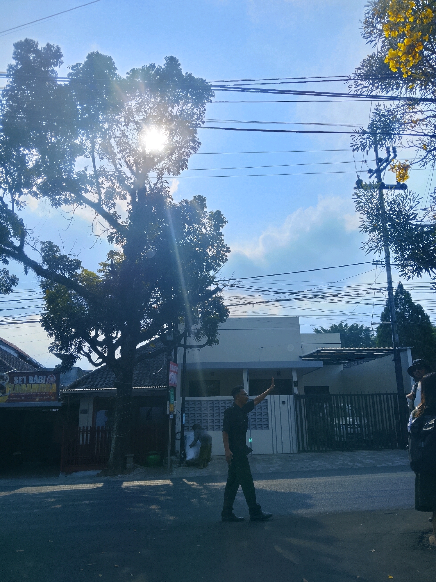 The image captures a street scene in Malang, Indonesia, under a bright, sunny sky with scattered clouds. A large tree with dense foliage dominates the left side of the frame, with the sun shining through its leaves, creating lens flare. Power lines are strung across the sky. On the right, a utility pole with yellow flowers is visible. In the foreground, a man in a dark uniform, possibly security or police, is walking on the road, raising his right hand as if signaling or greeting. Behind him, a modern white building with a fence and a parked car can be seen. To the left of the man, a building with a tiled roof and a sign reading "SE'I BABI FLORA MORA" is visible, suggesting a food establishment. Further to the right, the shoulders and heads of two individuals, one wearing a hat, are partially visible, indicating their presence on the sidewalk or street edge. The road surface shows some shadows from the trees and possibly debris. The overall impression is a typical urban street view in Malang during daylight hours.