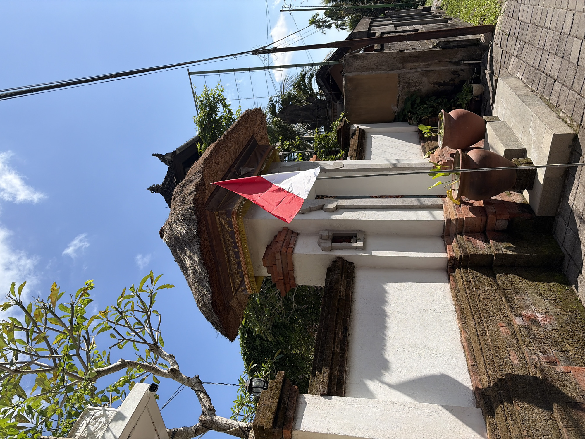 In a bright outdoor scene set in Ubud District, Indonesia, an Indonesian flag, a red and white banner, is prominently displayed on a pole in front of a traditional building entrance. The entrance features a thatched roof, red brick accents, and white stucco walls. Two large terracotta pots containing plants sit on a raised platform to the right of the entrance. The area is paved with brick. In the background, lush green foliage, trees, and power lines are visible against a blue sky with scattered clouds. The image is taken from a low angle, looking slightly upwards.
