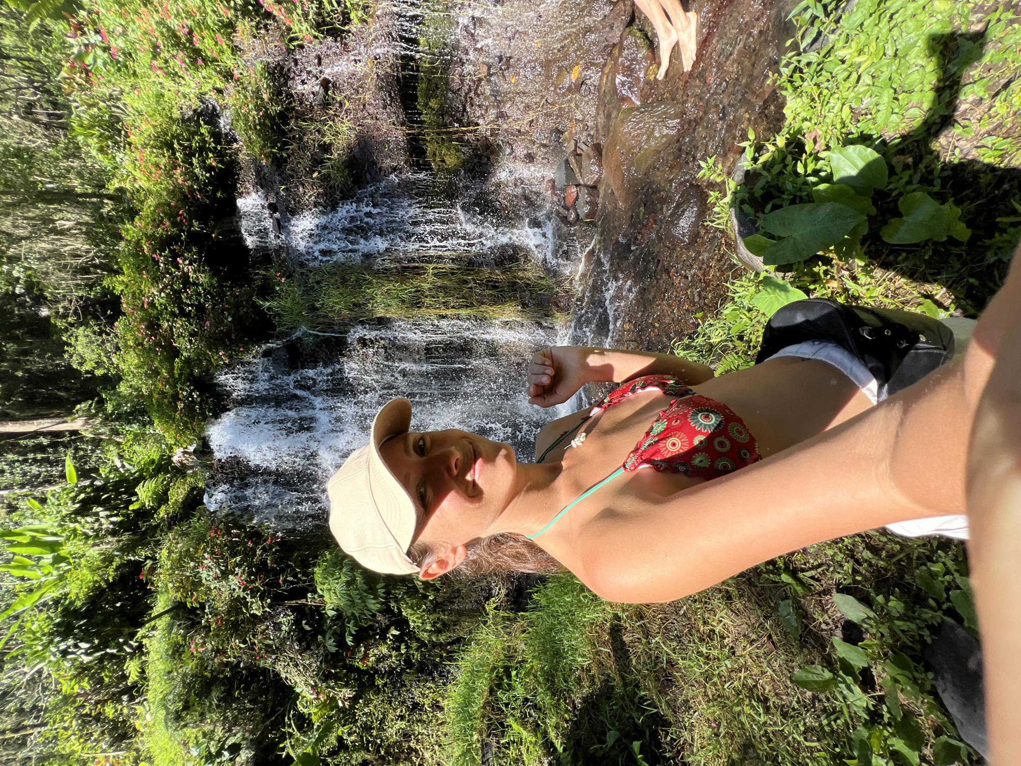 In Busungbiu, Indonesia, a young woman in a red and gold patterned bikini and a beige baseball cap smiles at the camera while sitting near a waterfall. Her left arm is extended, and her right arm is bent at the elbow. The waterfall flows over moss-covered rocks and is surrounded by lush green foliage, including ferns and small red flowers. The sun shines brightly, casting shadows on the grassy ground. In the background, two people's legs and feet are visible standing in the water. The woman appears relaxed and happy in the natural setting.