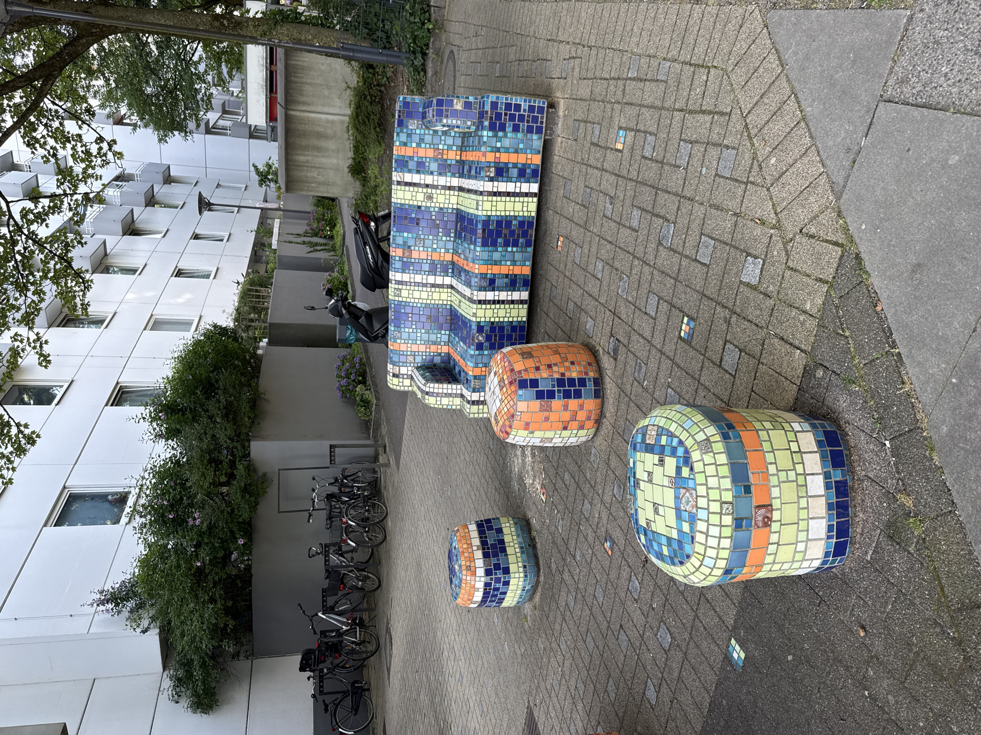 In Hamburg, Germany, a mosaic-covered outdoor seating area features a brightly colored sofa and three cylindrical stools. The furniture is made of ceramic tiles in various shades of blue, green, orange, and white, arranged in geometric patterns. The sofa has a striped design, while the stools have more random patterns. The seating area is located on a paved courtyard with a brick pattern. In the background, a modern apartment building with white facades and balconies stands behind lush green bushes and trees. Several bicycles are parked to the left of the building, and two scooters are visible near the entrance. The overall scene suggests a well-maintained urban public space with an artistic touch.
