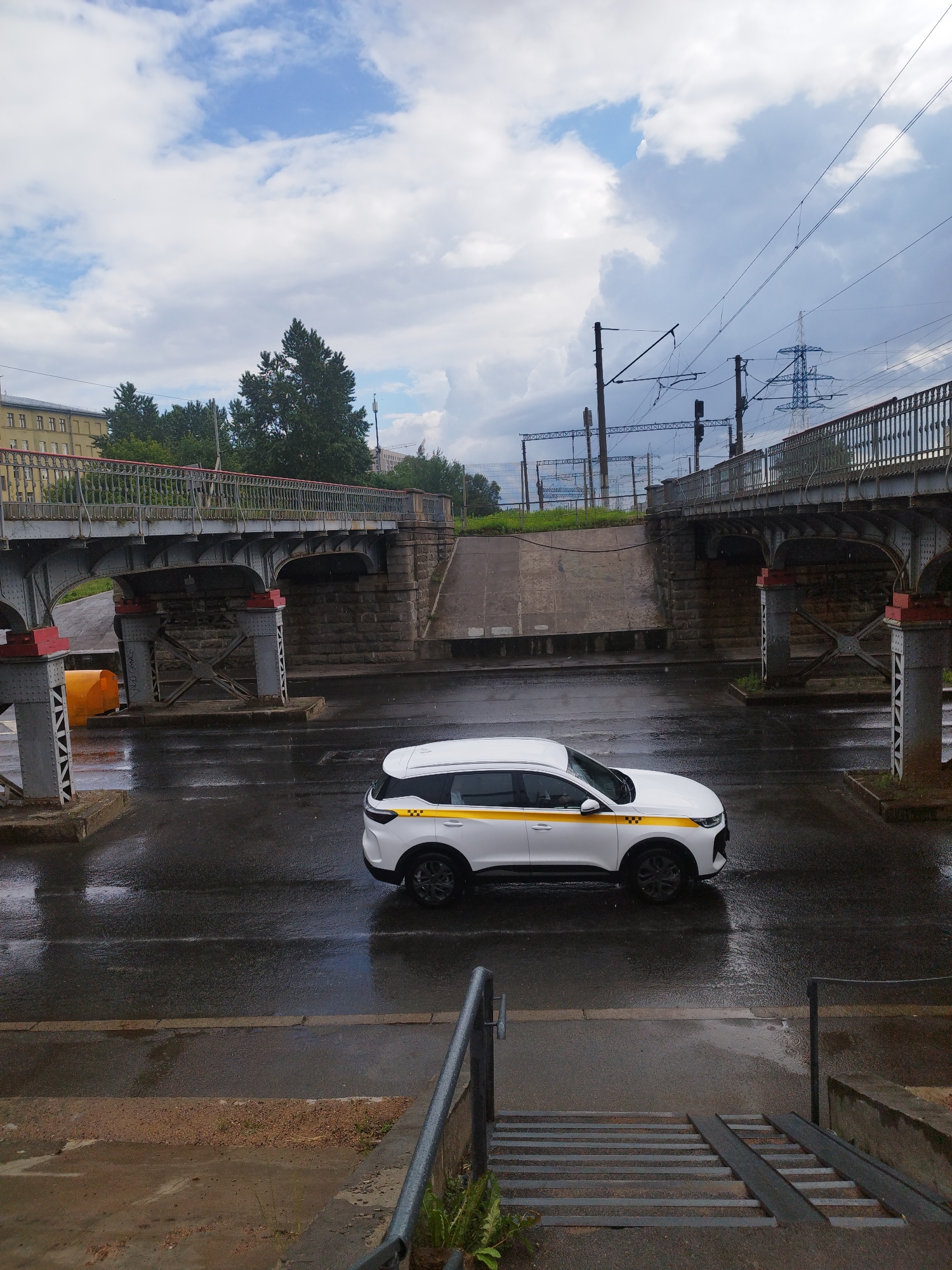A white taxi with a yellow stripe drives on a wet, asphalt road under an overcast sky. The taxi is a small SUV. In the background, a metal pedestrian bridge with red accents crosses over the road. Beyond the bridge, there are train tracks with overhead power lines and a tall, blue electrical tower. To the left of the bridge, a yellow object, possibly a traffic cone, is visible. The scene appears to be in an urban environment, possibly in St. Petersburg, Russia, given the architectural style of the bridge and the presence of an electrical tower common in Russian cities. The ground is wet, suggesting it may have recently rained or is currently raining lightly.