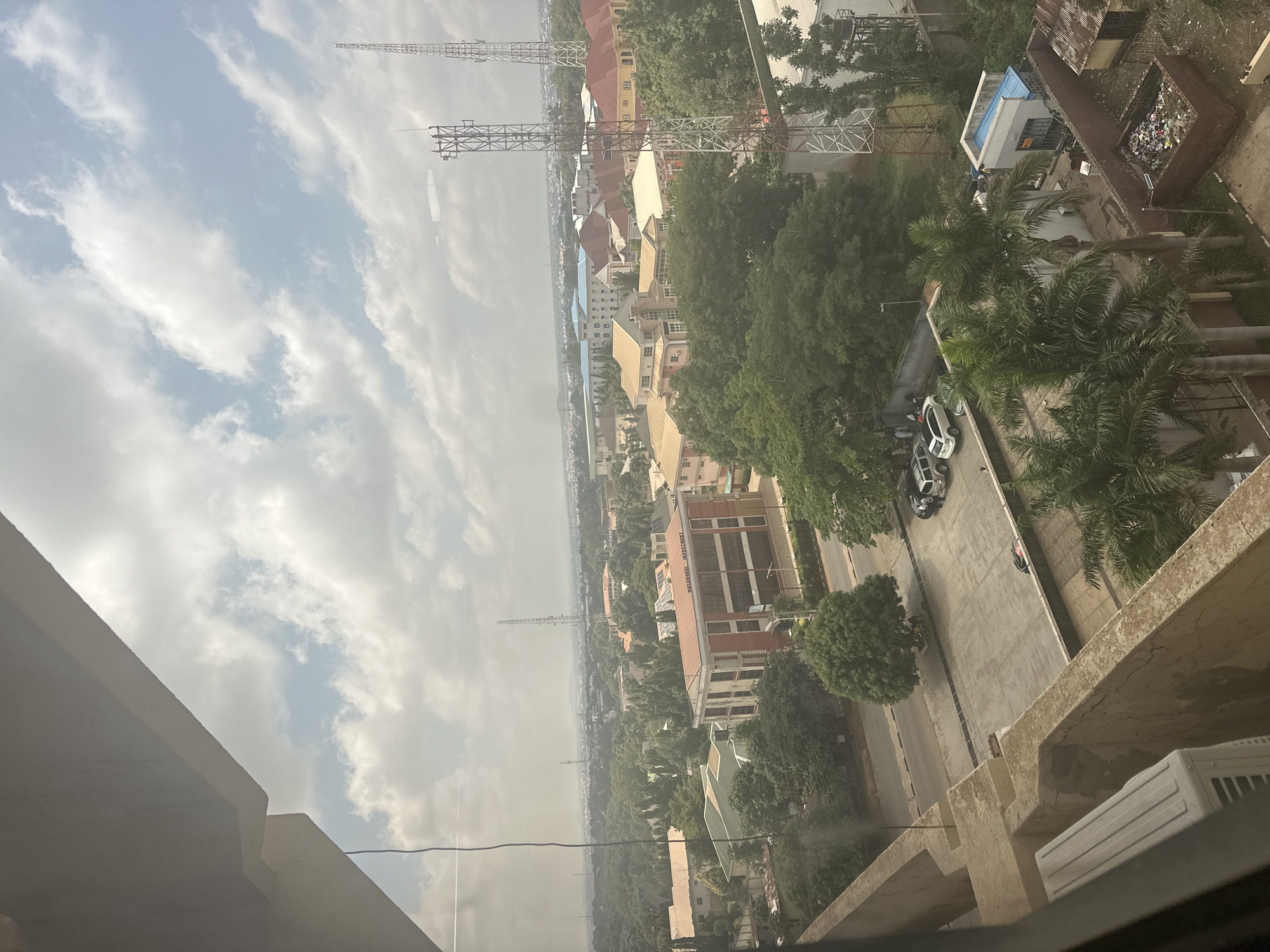 The image provides a high-angle view of an urban environment, likely in Shaba, Nigeria, under a partly cloudy sky. In the foreground, a concrete balcony edge juts into the frame, with a white air conditioning unit visible on its surface. The middle ground features a cityscape with numerous buildings of varying architectural styles, many topped with red or brown tiled roofs. Lush green trees are interspersed throughout the urban landscape, suggesting a well-treed area. Several telecommunications towers, characterized by their lattice metal structures, are prominent features in the scene, indicating a developed area with communication infrastructure. A road is visible in the middle distance, with parked cars, including at least two SUVs and a sedan. In the lower right portion of the image, a small building with a blue roof and a brick enclosure filled with trash are visible. The overall impression is one of a bustling city or town with a mix of residential and commercial structures, surrounded by abundant vegetation.