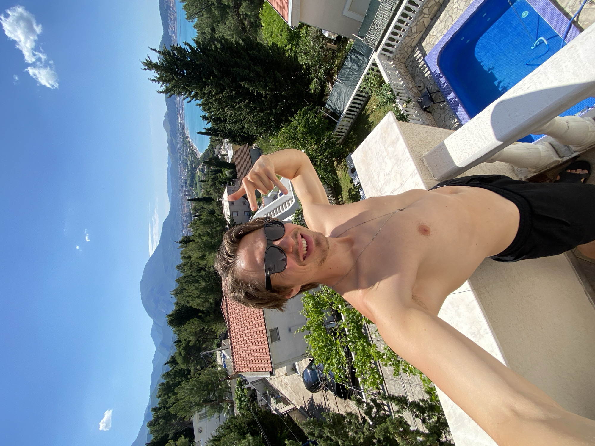 A shirtless man with sunglasses stands on a balcony, making a hand gesture. The balcony overlooks houses, trees, and a distant coastline and mountains. To the right is a pool. The location appears to be in a mountainous area, likely the city of Šušanj, Montenegro.