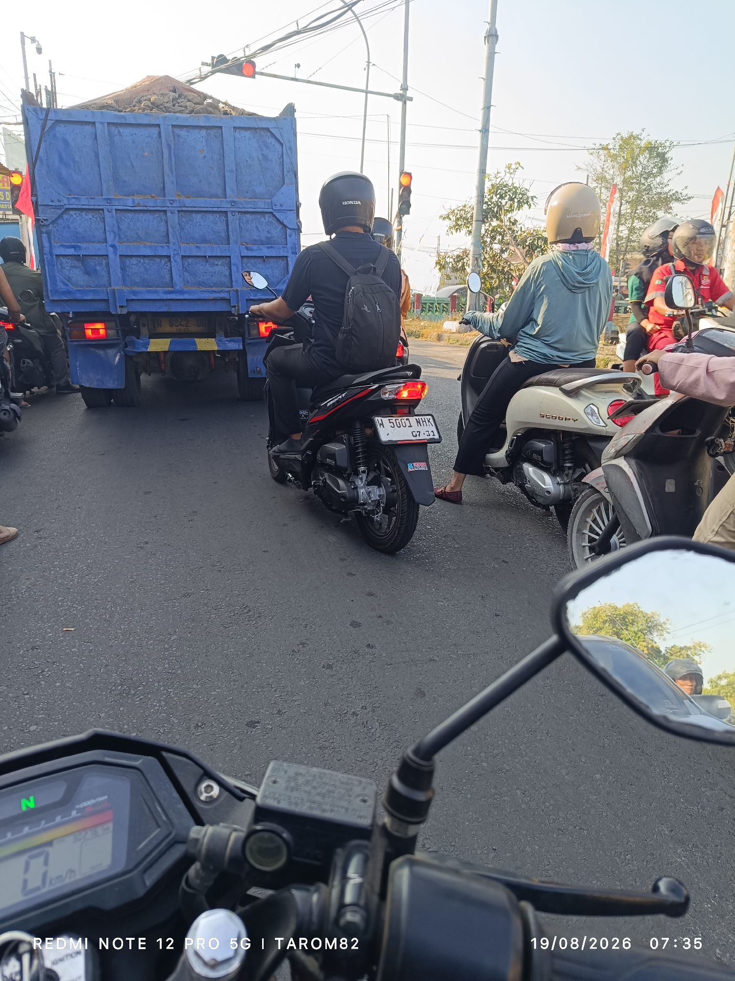 This image, taken in Sukodono, Indonesia on August 19, 2026, at 07:35, depicts a traffic scene. A large blue dump truck filled with what appears to be dirt or rubble is stopped at an intersection, indicated by a red traffic light. Several motorcycles are also stopped behind the truck. One motorcycle, a black Honda Beat with license plate W 5001 NHK, is prominent in the foreground on the left. Its rider, wearing a black helmet and backpack, is looking forward. To the right of the truck, another motorcycle, a white Honda Scoopy, is stopped. Its rider, wearing a light blue hoodie and a beige helmet, is also facing forward. Further to the right, another motorcycle with a rider wearing a red shirt and helmet is visible. The environment appears to be a paved road with some vegetation and utility poles in the background. The sky is clear and bright, suggesting it is daytime. The perspective suggests the photo is taken from another motorcycle.
