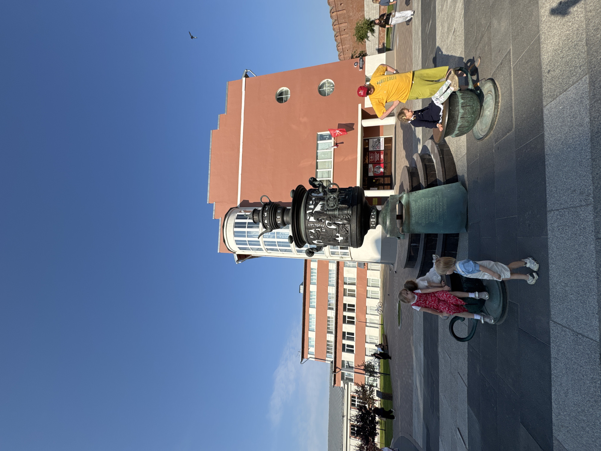 The image depicts a sunny day in Tula, Russia, likely in a public square given the presence of a large, ornate samovar monument. In the foreground, a group of children are interacting with the base of the samovar, which appears to be designed for seating or climbing. One young girl in a red dress sits on a portion of the monument, while a younger child in a blue outfit stands nearby. Another child, in a dark dress, is seated on a smaller, cup-shaped element of the monument. An adult man, wearing a yellow t-shirt and green trousers, stands behind the children, observing them.
In the background, a distinctive building with a rounded, glass-enclosed section and a reddish-orange facade dominates the scene. Several other buildings, appearing to be institutional or administrative, are visible to the left, with a brick wall visible to the far right. The sky is a clear, vibrant blue, and a bird is captured in flight high above. The overall atmosphere suggests a casual outdoor gathering or tourist activity. The samovar monument is a prominent cultural symbol of Tula, a city historically renowned for its samovar production.