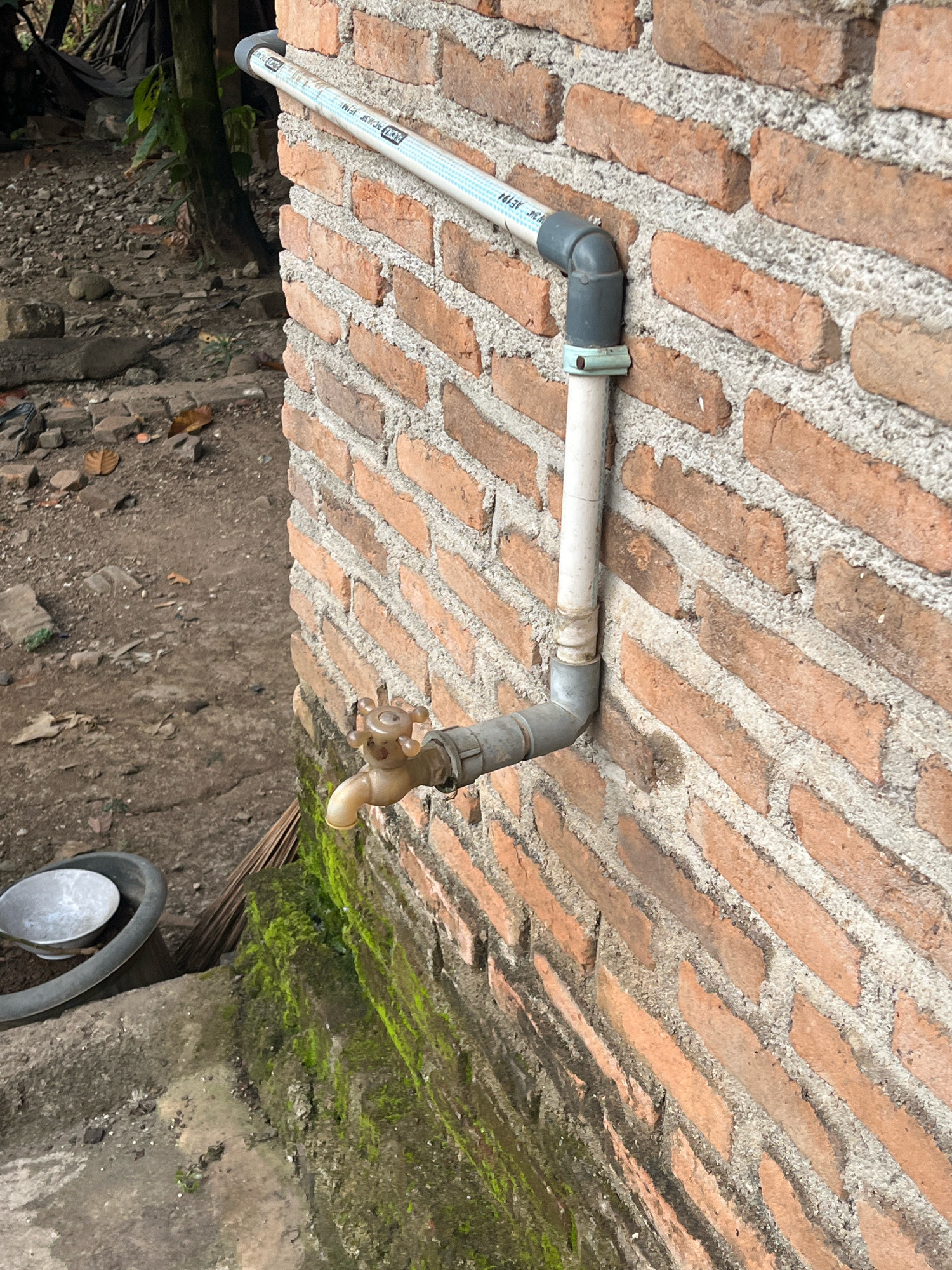 The image depicts an outdoor scene in Kebumen, Indonesia, focusing on a water faucet attached to a brick wall. The faucet is made of a light-colored material and has a four-pronged handle. It is connected to PVC piping that runs horizontally along the brick wall and then vertically downwards. The brick wall is constructed from reddish-brown bricks with gray mortar. Below the faucet, the wall is partially covered in green moss. To the left of the wall, there is a patch of dirt with scattered debris, including small stones and fallen leaves. A dark gray plastic basin and a broom are also visible on the ground near the wall. The overall impression is of a utilitarian outdoor area, possibly in a residential setting.