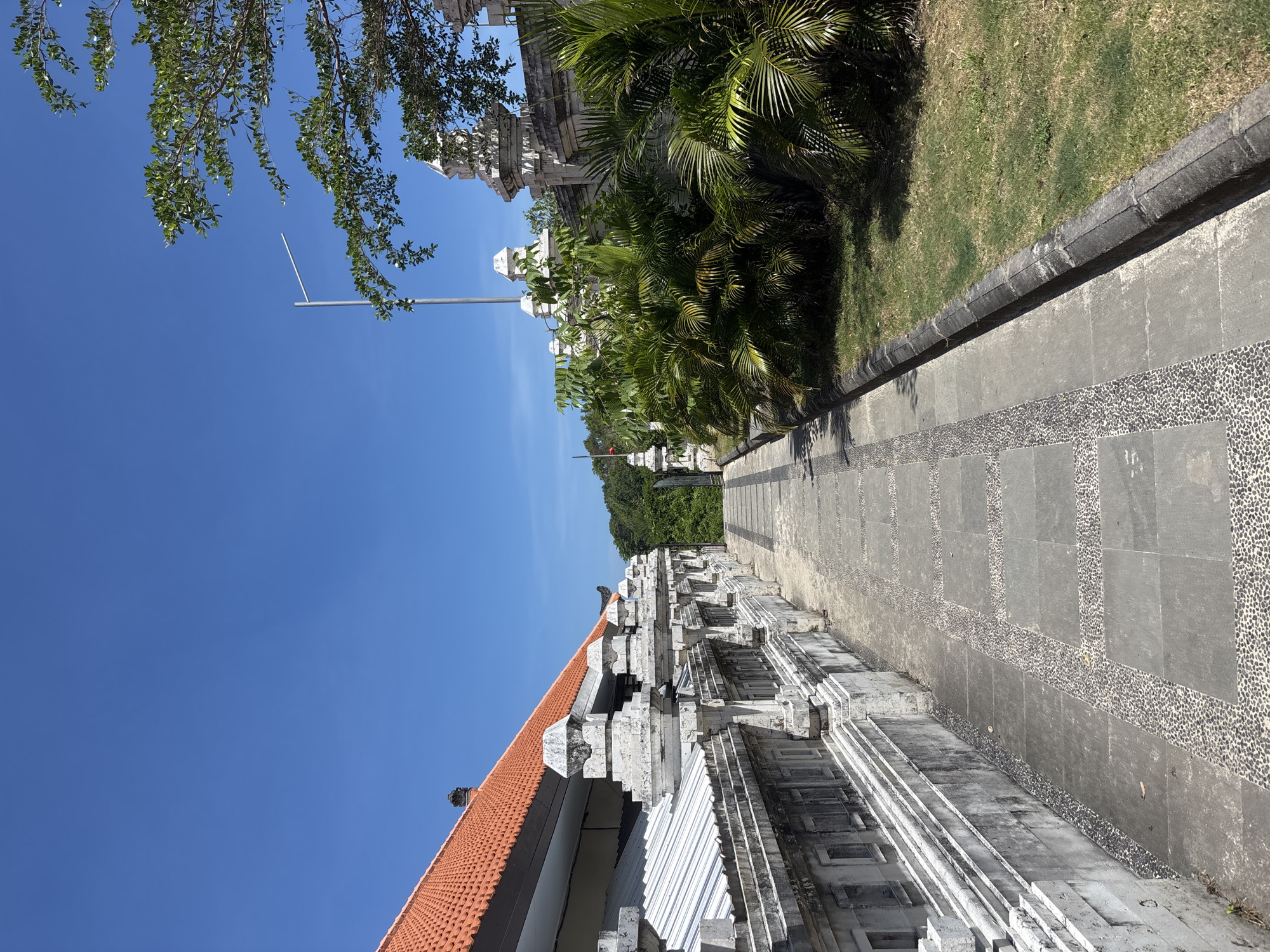 The image depicts a pathway alongside a wall or structure in Kuta Selatan, Indonesia. The path is constructed with square stone slabs and interspersed with sections of small, rounded stones. A low curb separates the path from a patch of green grass and various tropical plants, including palm trees. A tall, white structure with multiple tiers and a red tile roof runs along the opposite side of the path. The sky is clear and blue, and a few tree branches are visible in the upper left corner of the frame. A metal pole stands tall in the distance. The overall impression is of a well-maintained outdoor space, likely within a park or temple complex.