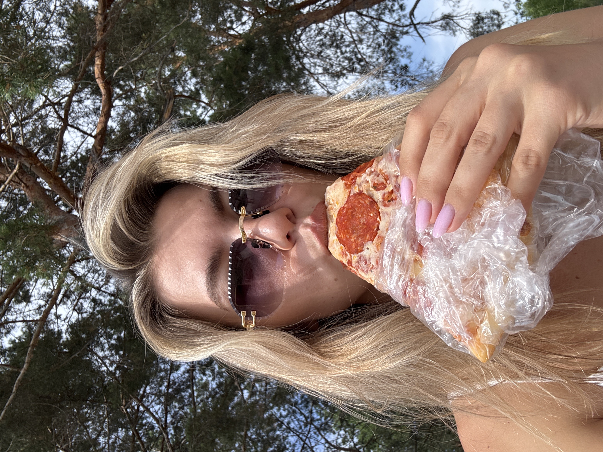 A woman with blonde hair and pink fingernails is taking a bite of a slice of pepperoni pizza. She is wearing large sunglasses with dark lenses and gold frames. The pizza is wrapped in clear plastic. The background is blurred and shows green trees and a bright sky, suggesting an outdoor setting during the daytime. The woman's expression is neutral as she bites into the pizza.