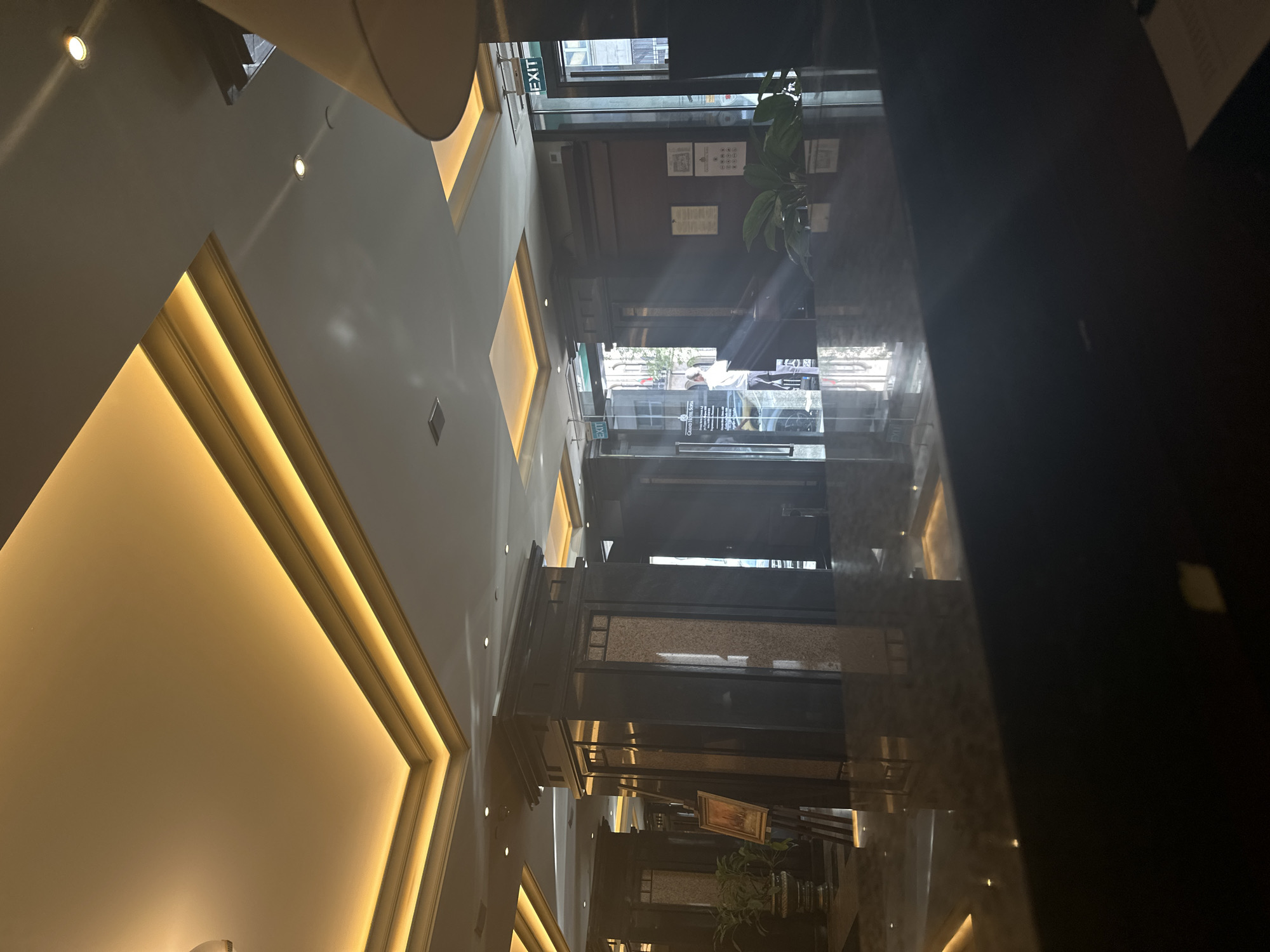 The image depicts the interior of a building, likely a hotel or office, with a multi-story atrium. The perspective is looking upwards. There is a false ceiling with recessed lighting in a geometric design. The atrium features dark-colored pillars or support structures that extend upwards, with some decorated with stone-like panels. The presence of an "EXIT" sign suggests that the building is located somewhere in Bulgaria. The location is Sofia, Bulgaria.