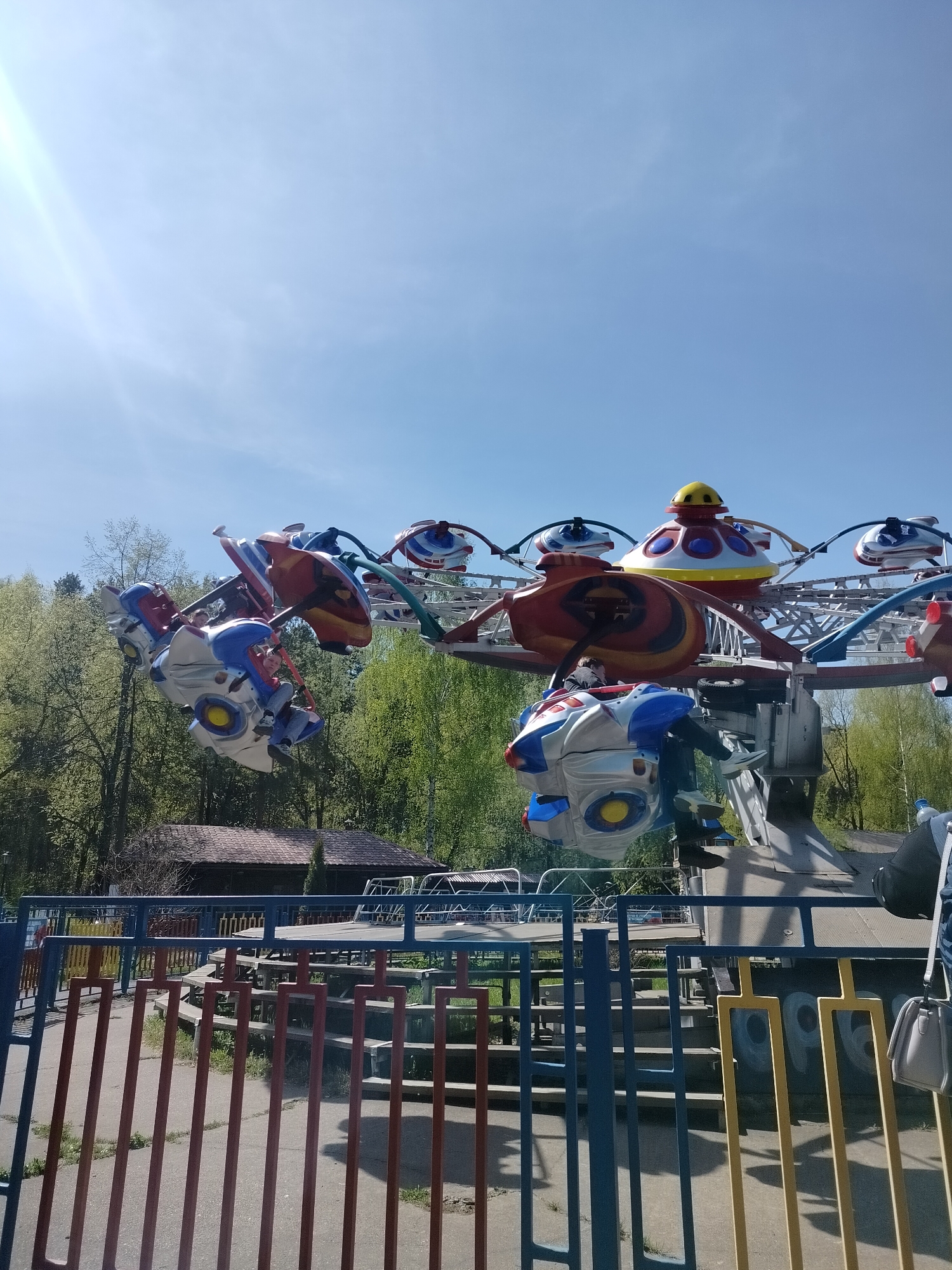 The image captures a fairground ride in motion, likely located in the city of Chaikovsky, Russia. The ride features several spaceship-themed cars rotating around a central axis. Occupants are visible within the cars. The ride is enclosed by a safety fence painted in multiple colors. Trees are visible in the background, indicating the park or outdoor setting. The sky is clear and blue, suggesting a sunny day. A small building with a tiled roof can be seen in the background. A purse on a person's shoulder is visible near the bottom right of the image.