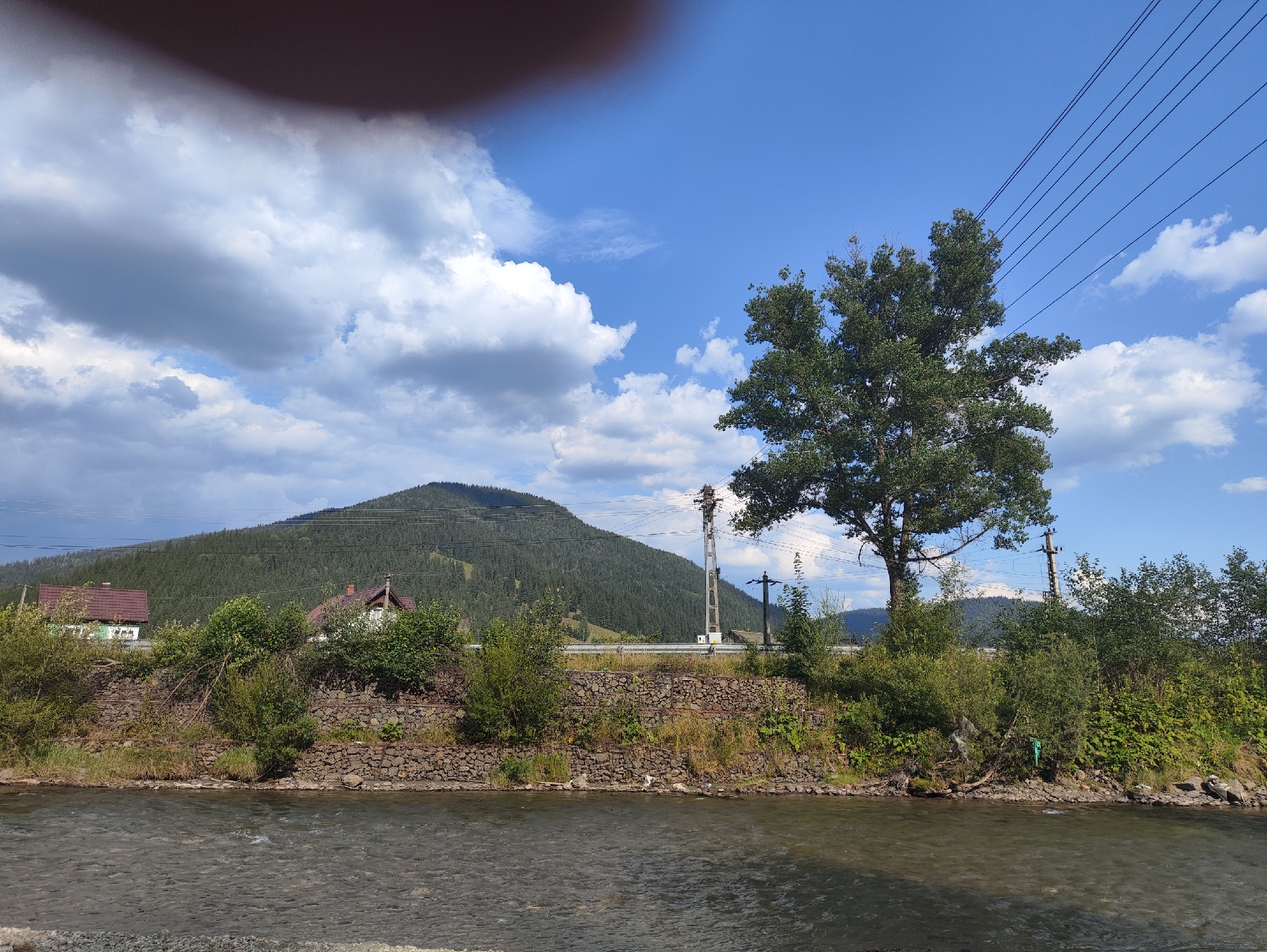 The image depicts a serene natural landscape, possibly in the vicinity of Botoș, Romania, characterized by a clear blue sky with scattered white clouds. A river flows gently in the foreground, its surface rippling with a subtle current. The riverbank is lined with dense green foliage and trees, with a stone embankment reinforcing the terrain.
In the middle ground, a verdant mountain, covered in a thick carpet of coniferous trees, rises majestically. A few houses with reddish-brown roofs are nestled at the base of the mountain, suggesting a rural or semi-rural settlement. Power lines stretch across the scene, supported by utility poles, indicating the presence of infrastructure. A large, leafy tree stands prominently to the right of the frame, its branches reaching towards the sky. The overall impression is one of natural beauty and tranquility, with a subtle human presence.