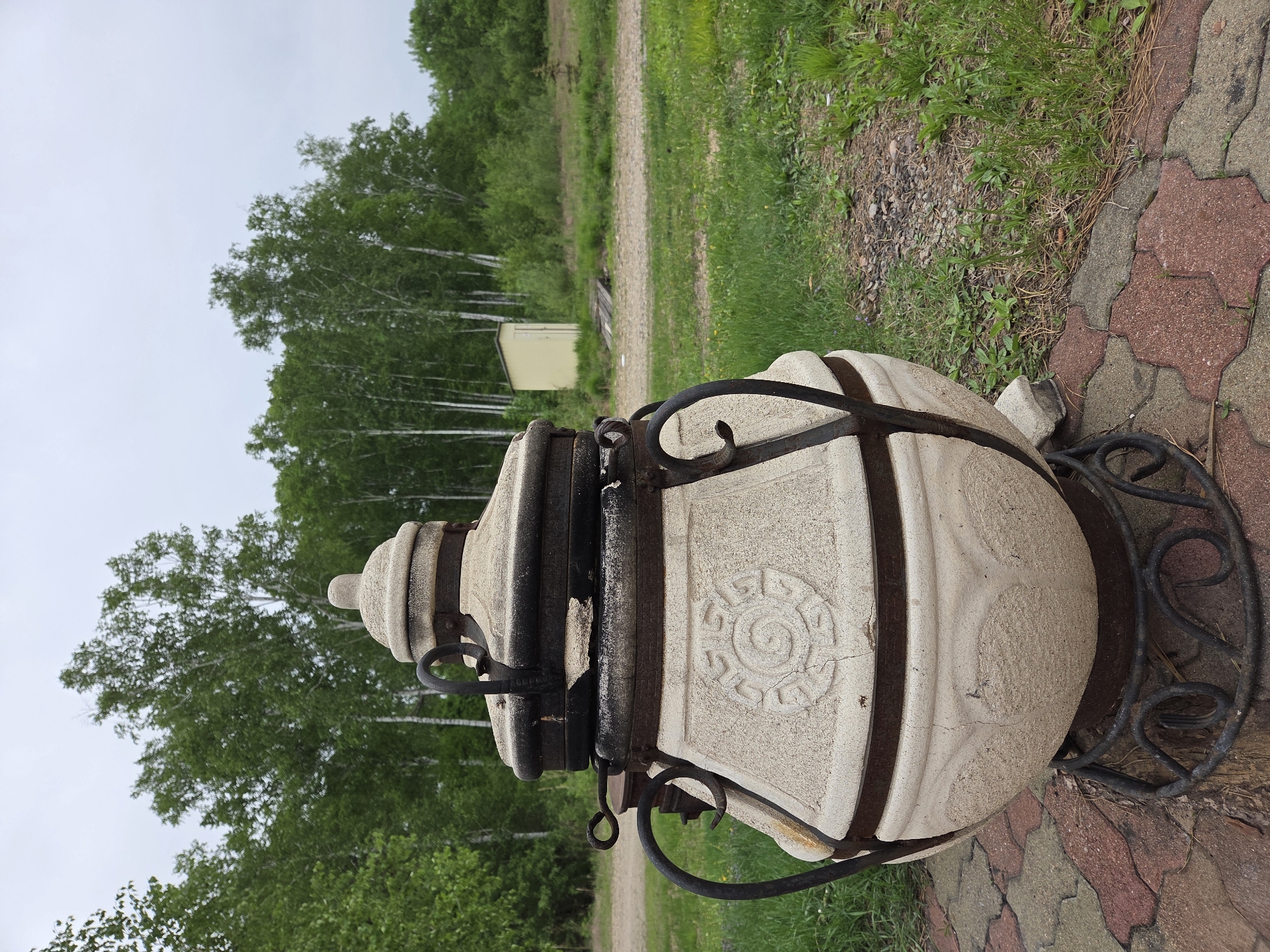 A stone tandoor oven sits outdoors on a wrought-iron stand. The oven is light-colored, possibly white or off-white, with decorative engravings and horizontal metal bands around it. The stand has a decorative, ornate design. The oven and stand are located on a paved area made of red and grey interlocking stones. Behind the oven is a grassy area with trees and a dirt path. A small, light-colored building is visible in the distance amidst the trees. The scene appears to be in a natural or park-like setting. The location is stated to be Birobidzhan, Russia, though the image itself does not explicitly confirm this.