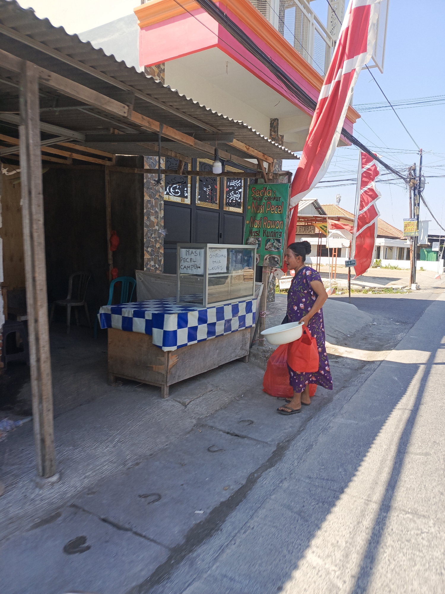 The image captures a street-side food stall in Dukuhtengah, Indonesia. A woman wearing a purple floral dress stands in front of the stall, holding a white bowl in her left hand and two red plastic bags in her right. The stall itself is covered by a corrugated metal roof supported by wooden beams. In front of the stall, a wooden counter is covered with a blue and white checkered tablecloth. A glass display case sits on top of the counter, containing food items. A green sign above the display lists menu items: "Sedia: Nasi Campur, Nasi Pecel, Nasi Rawon, Nasi Kuning" and "ONIONG MIE LODEH". Two chairs are visible inside the covered area of the stall. Red flags are prominently displayed, one large one on a pole to the right of the stall and another smaller one behind it. The street pavement is visible in the foreground, with shadows cast by the stall and flags. The background shows residential buildings and utility poles against a clear blue sky.