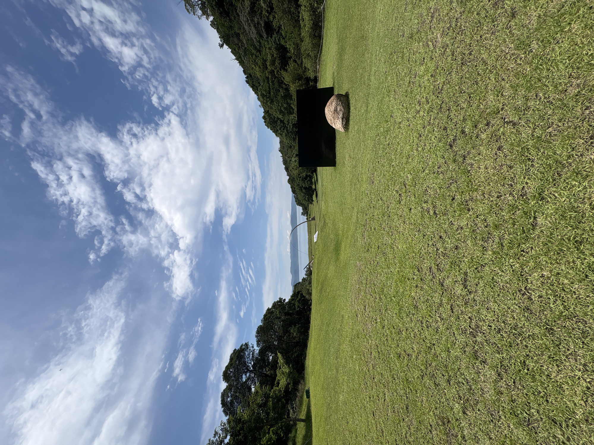 The image shows a wide, green lawn in front of a body of water. In the foreground, there is a large, rounded boulder placed in front of a black, rectangular structure that appears to be an art installation. The lawn slopes gently towards the water, which is calm and reflects the blue sky with scattered white clouds. Behind the lawn, a dense forest covers a hillside that meets the water. In the distance, across the water, there are faint outlines of mountains. The sky is a clear blue with wispy clouds. The scene is likely part of the art installations on Naoshima Island, known for its contemporary art museums and outdoor sculptures.