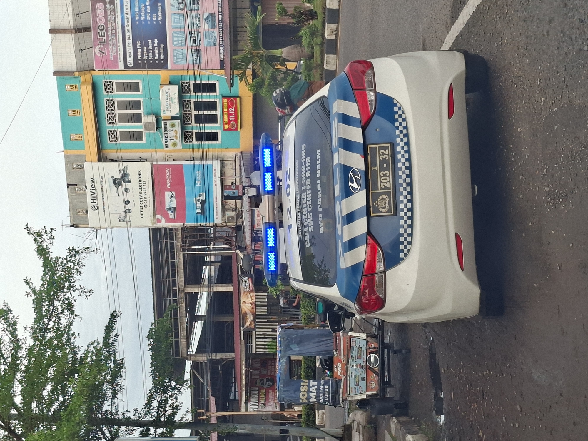 The image shows a white police car with blue flashing lights parked on a street in Lhokseumawe, Indonesia. The car has the license plate "I 203 - 32". In the background, there are several multi-story buildings with various advertisements and signs, including "HiView", "Optus CCTV Elektronik", "Hikvision", and "LEGOS". A small, two-wheeled cart is parked next to the police car. A person wearing a helmet is partially visible on a motorcycle near the buildings. The weather appears overcast.