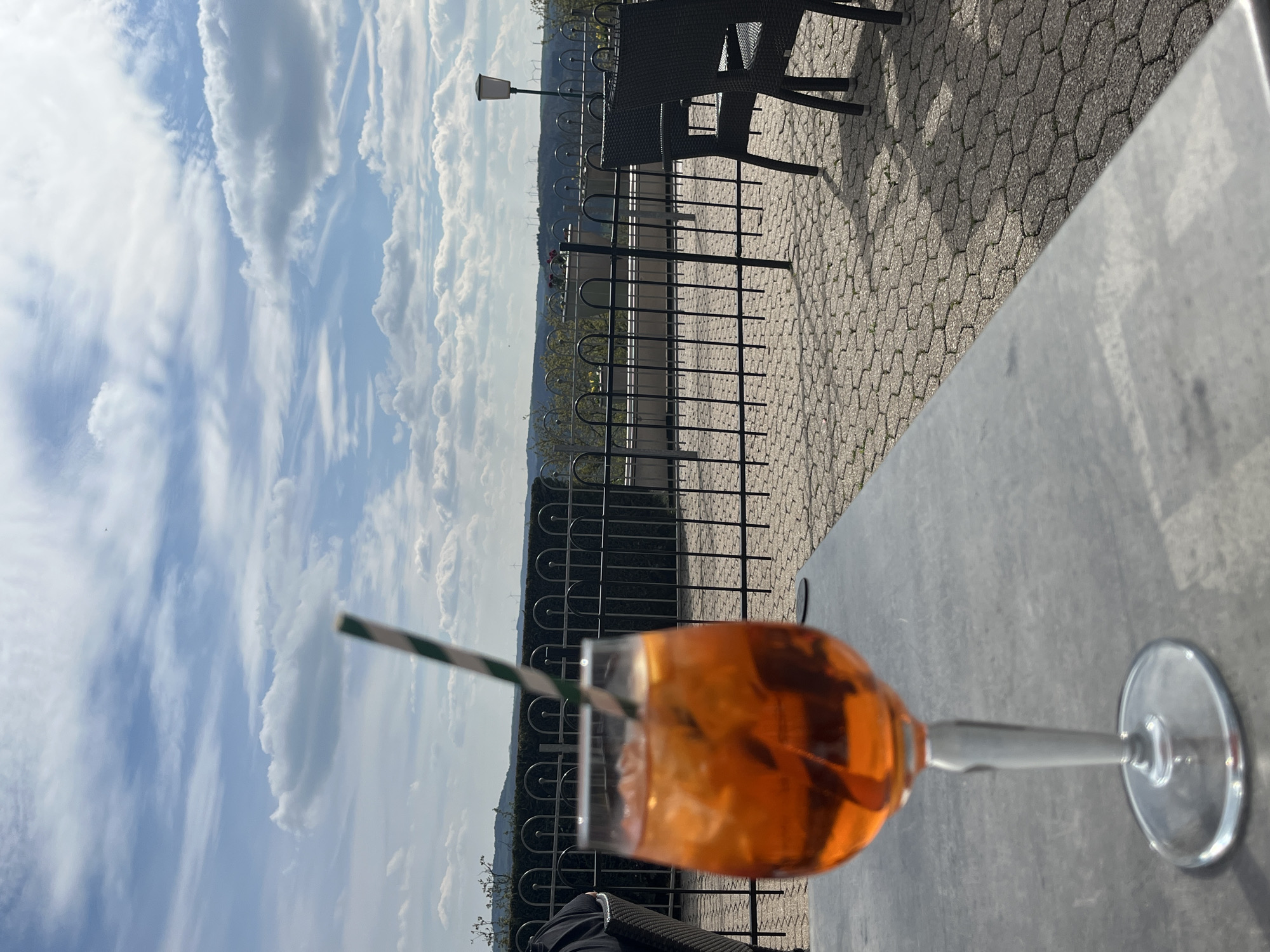 A cocktail glass filled with an orange liquid is prominently displayed on a grey table surface, with a green and white striped straw protruding from the glass. Behind the glass is a black metal railing. Beyond the railing, a background landscape includes trees, possibly a river valley, and a sky filled with clouds. To the upper right, black patio furniture sits on a hexagonal stone-paved surface. The image appears to be taken from a slightly elevated perspective. Based on the presence of a river valley and patio furniture, the location is likely an outdoor seating area in Saarburg, Germany, given the prompt specified the location.