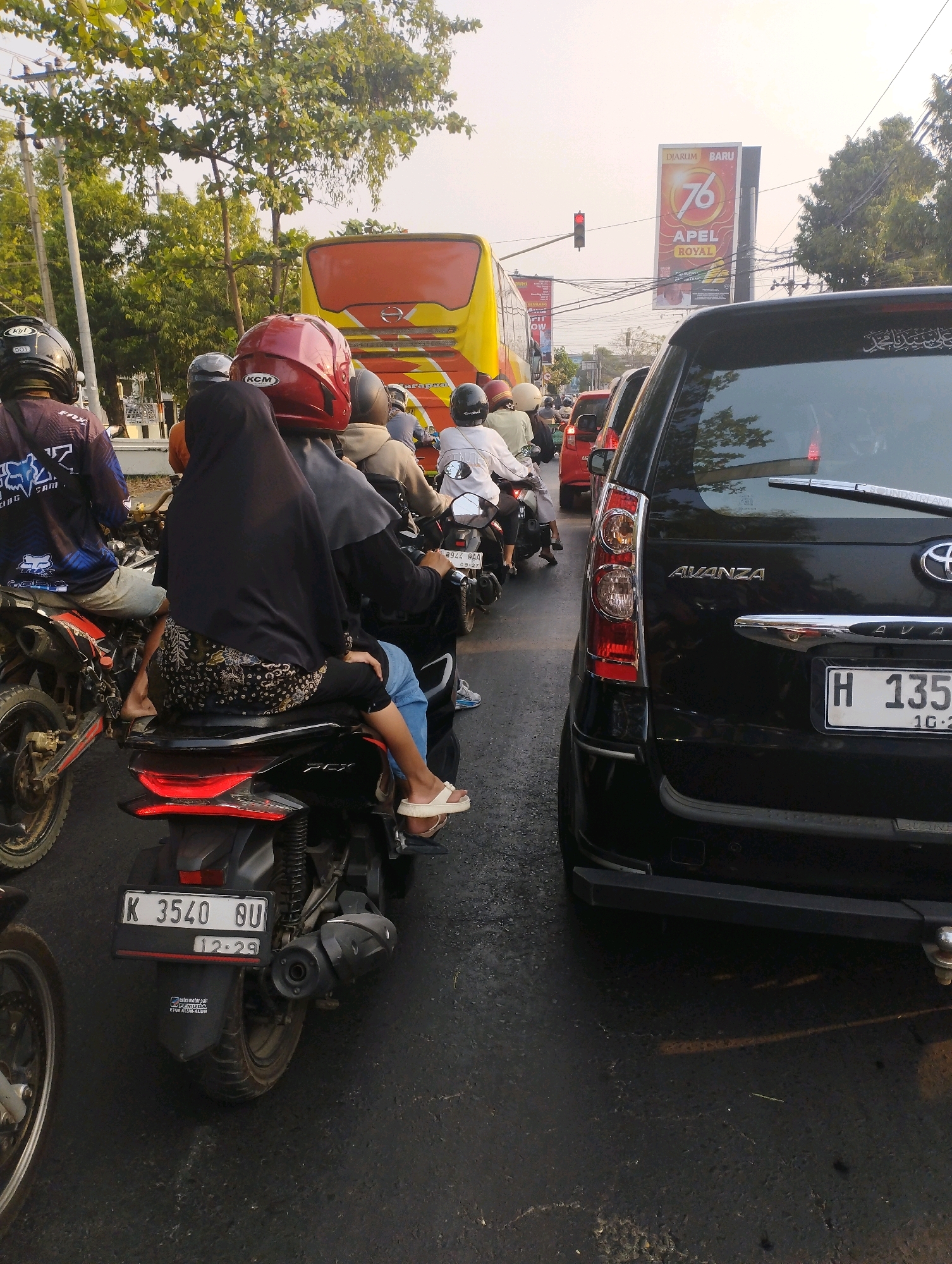 The image depicts a traffic jam on a street in Kecamatan Pati, Indonesia. A black Toyota Avanza is the primary subject, positioned in the foreground and facing away from the camera. Behind it, numerous motorcycles and a yellow bus are visible, also stopped in traffic. The scene appears to be on a paved road with trees and utility poles lining the sides. Several people are on the motorcycles, some wearing helmets. A billboard with the text "DIARUM BARU 76 APEL ROYAL" is visible in the background. The lighting suggests it is daytime, possibly late afternoon given the quality of light. The overall impression is one of heavy traffic congestion in an urban or semi-urban environment.