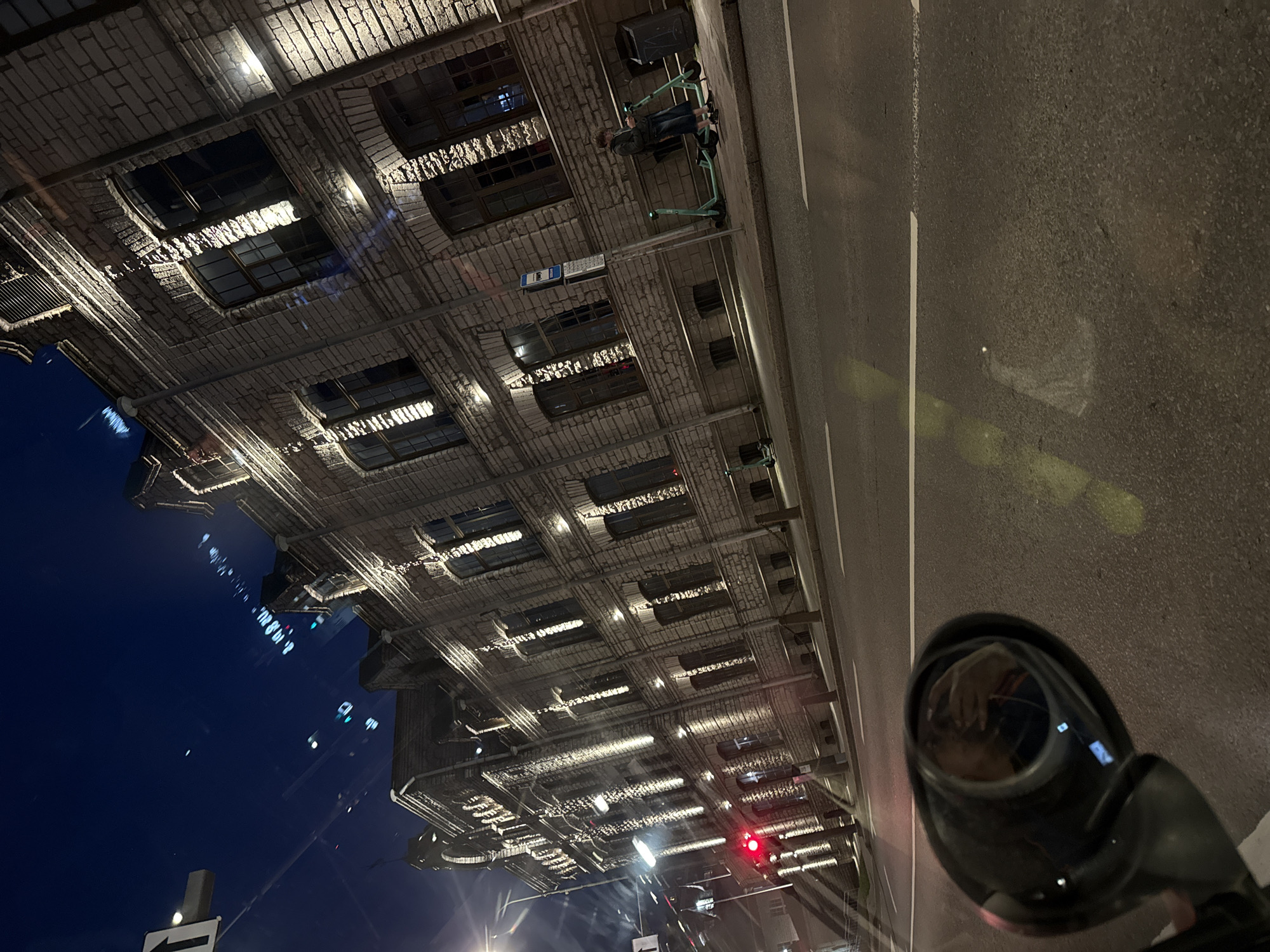 The image captures a street view in Tallinn, Estonia at night. The scene features a multi-story, stone-clad building with numerous windows, illuminated by exterior lighting. In the foreground, a black rearview mirror reflects a person holding a phone. The street is paved with asphalt, marked with white lines. On the sidewalk, several electric scooters are parked. A traffic light is visible, showing a red signal. The sky is dark blue, suggesting nighttime.