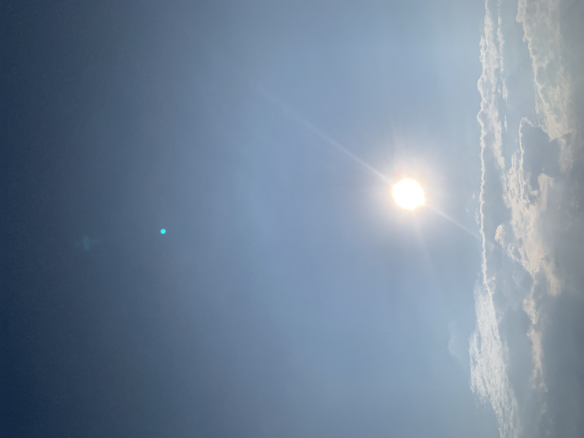 The image shows a bright sun low in the sky, casting rays of light across a pale blue sky. Below the sun are scattered white clouds, some appearing fluffy and others more wispy. The scene appears to be a daytime photograph taken from an outdoor perspective. No specific landmarks or elements indicating the city of Kolomyia, Ukraine are visible within the frame. The image focuses on the sky, sun, and clouds.
