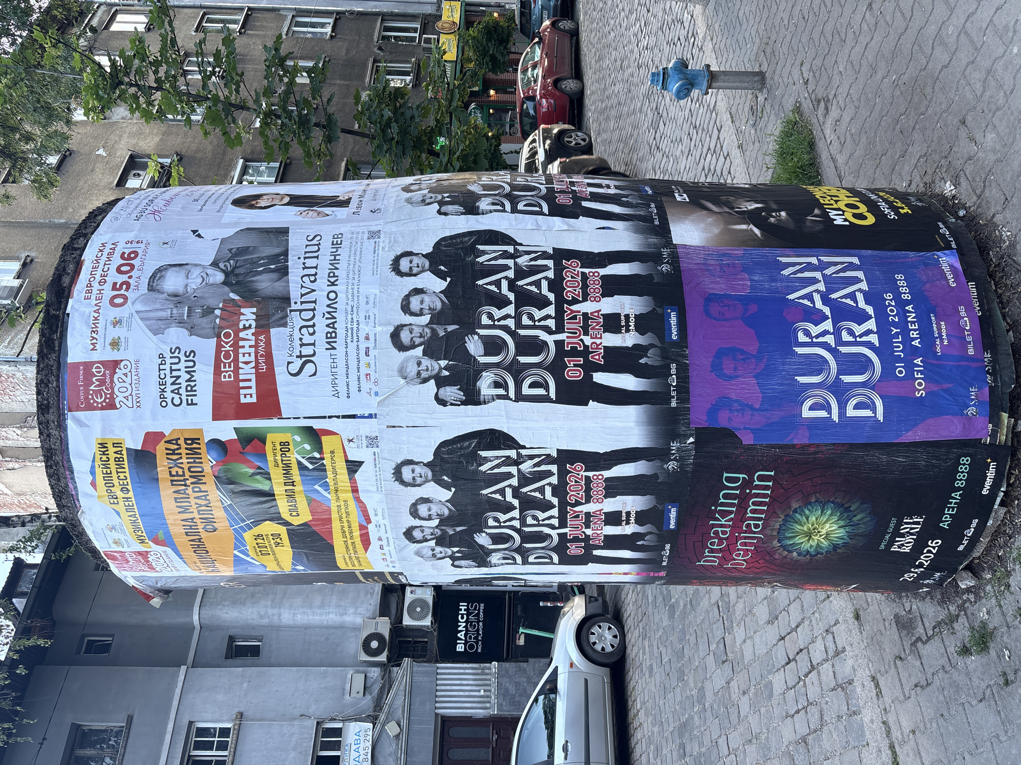The image depicts a cylindrical advertising column in Sofia, Bulgaria, plastered with various posters. The column is located on a sidewalk near buildings with visible air conditioning units and parked cars, including a white Bianchi Origins delivery van. A blue fire hydrant is visible on the sidewalk near the column. The posters advertise musical events, including concerts by Duran Duran (01 July 2026, Arena 8888, Sofia), Breaking Benjamin (29.6.2026, Арена 8888), the European Musical Festival (05.06, Hall Bulgaria). Additional posters promote the "National Youth Philharmonic" (07.07.26, Central Military Club). A "Stradivarius" concert with violinist Vesko Eshkenazi, conductor Ivaylo Krinchev, and other musicians is advertised. The surrounding environment suggests an urban setting with a mix of residential and commercial activity.