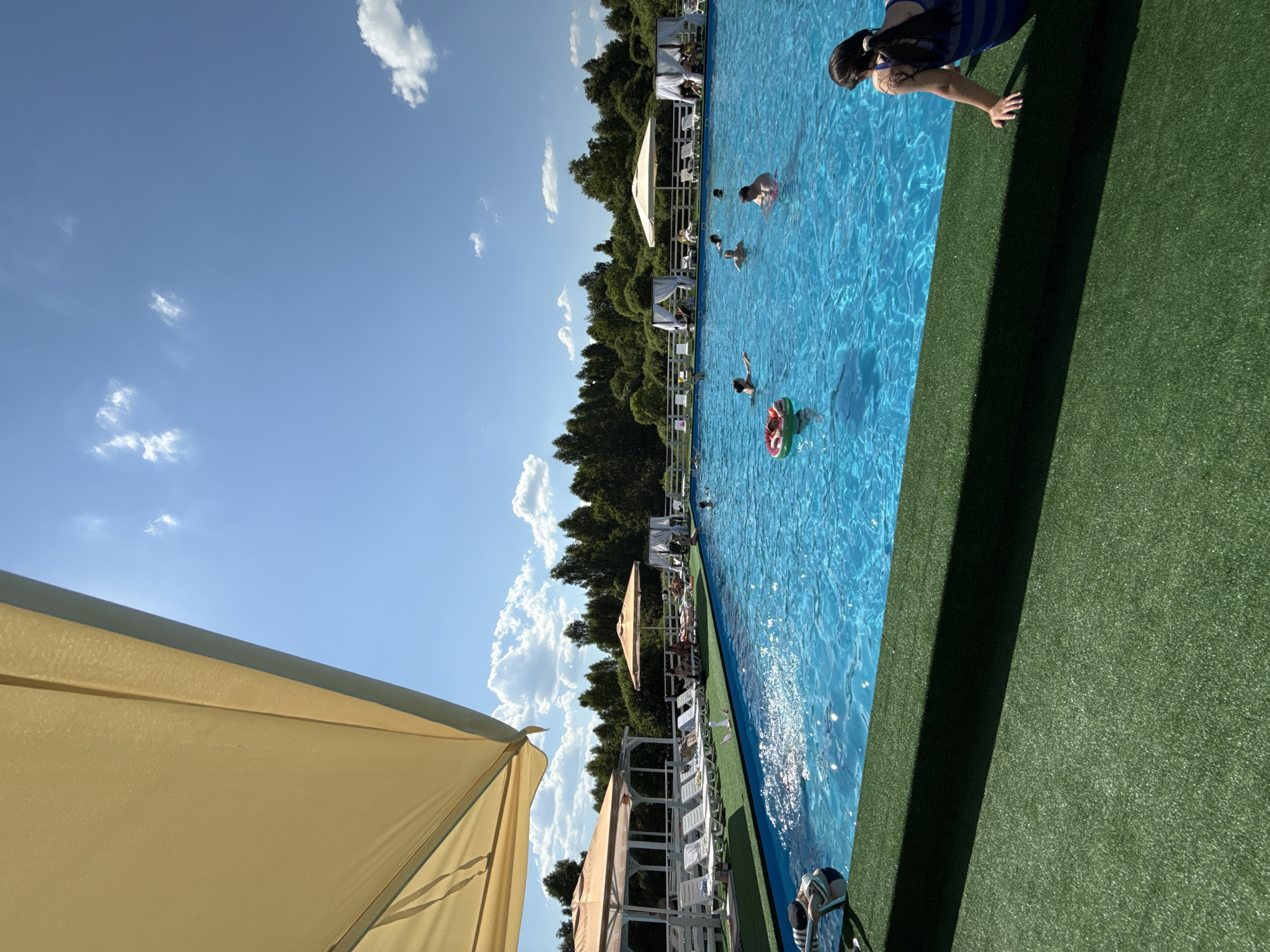 This is a daytime, eye-level, outdoor, wide shot of a swimming pool area. The pool is a bright blue and filled with people swimming and wading. There are lounge chairs and umbrellas surrounding the pool, and lush green trees in the background. The scene appears to be located at a resort or recreational facility in the village of Koloksha, Russia. The water is clear and sparkling, and the sky is a brilliant blue with a few fluffy white clouds. The overall atmosphere is one of leisure and relaxation on a warm, sunny day.