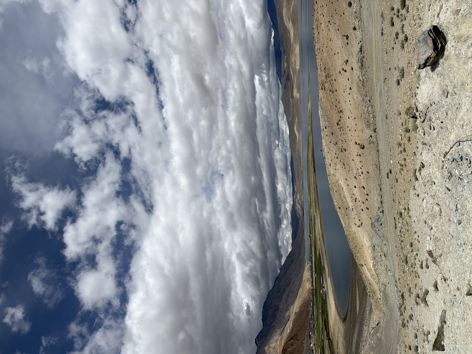 This is a vertical, eye-level, full shot of a vast, arid landscape with a large body of water and mountains under a dramatic sky, likely taken in Karzok, India.
In the foreground, the ground is covered in dry, sandy earth with scattered rocks and small green shrubs. A larger, dark, flat rock rests on the right side of the foreground. A dirt path winds through this area, leading towards the middle ground.
The middle ground features a wide, tranquil lake with calm, dark blue water. The shoreline is a mix of sandy areas and dry earth, with some green vegetation visible on the far side of the lake. Beyond the lake, rolling hills and mountains dominate the background, their slopes a mixture of brown, tan, and some hints of green. The peaks of the mountains are softened by distance and atmospheric haze.
A small village or settlement with buildings and green agricultural fields is nestled along the left bank of the lake, at the base of the mountains. The architecture appears traditional, with a cluster of white buildings and some red roofs.
The sky is a prominent feature, filled with a dramatic mix of dark, heavy storm clouds and patches of bright blue sky. The contrast between the dark clouds and the clear blue creates a sense of depth and impending weather change. Sunlight breaks through in places, illuminating parts of the landscape and the clouds.
There are no people prominently visible in the image, though there may be very small figures on the distant shore of the lake. The scene is one of natural grandeur and vastness, characteristic of high-altitude desert regions.